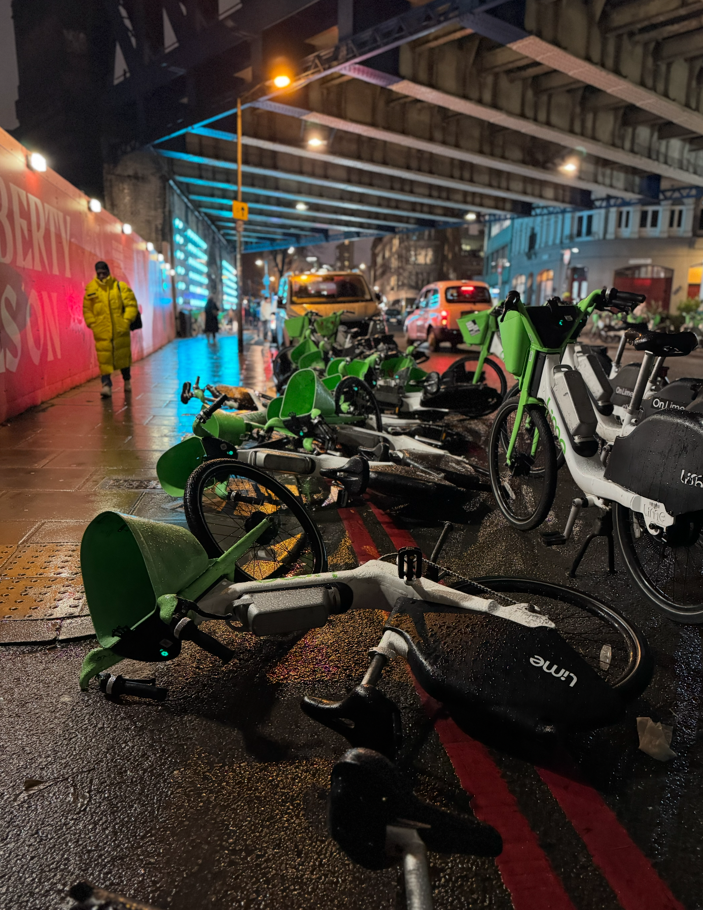 Fallen and overturned green and black bikes on a rainy city sidewalk under an overpass, with a person in a yellow coat walking by and yellow and white cars in the background.
