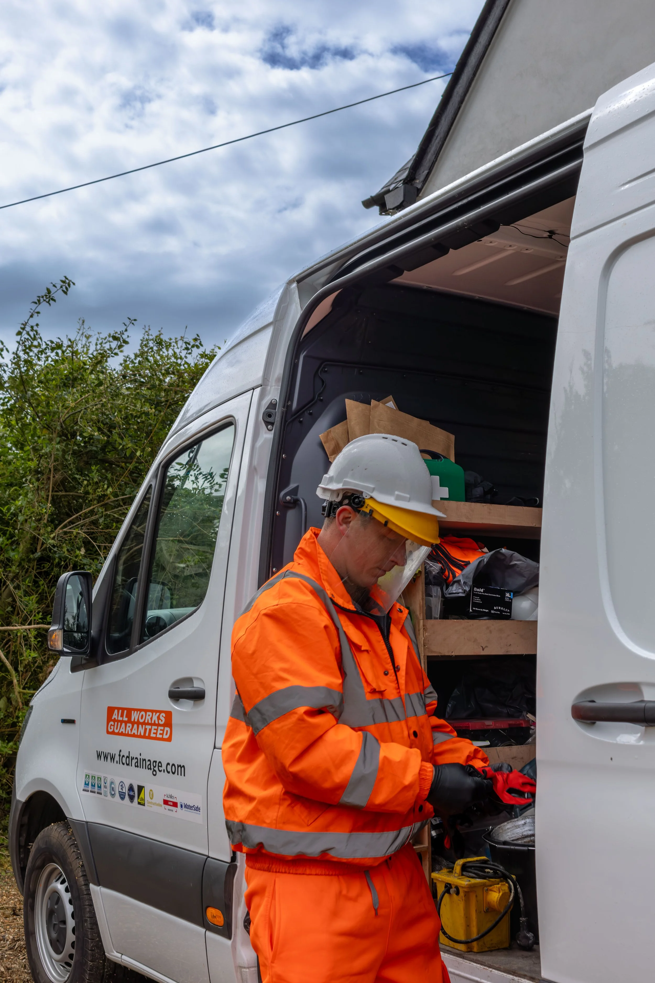 A worker in an orange safety suit and yellow helmet inside an open service van, with tools and equipment, with a cloudy sky and greenery in the background.