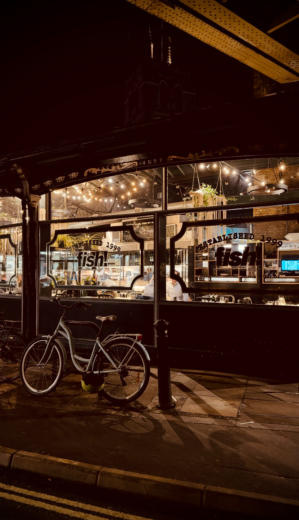 Nighttime view of a restaurant with string lights inside, large windows, and a bicycle parked outside on the sidewalk.