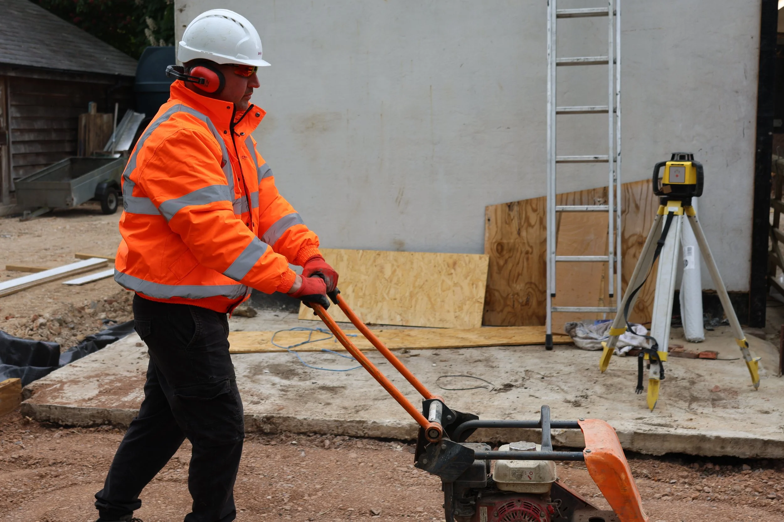 Construction worker in orange safety jacket and white hard hat operating a small compaction machine on a construction site.