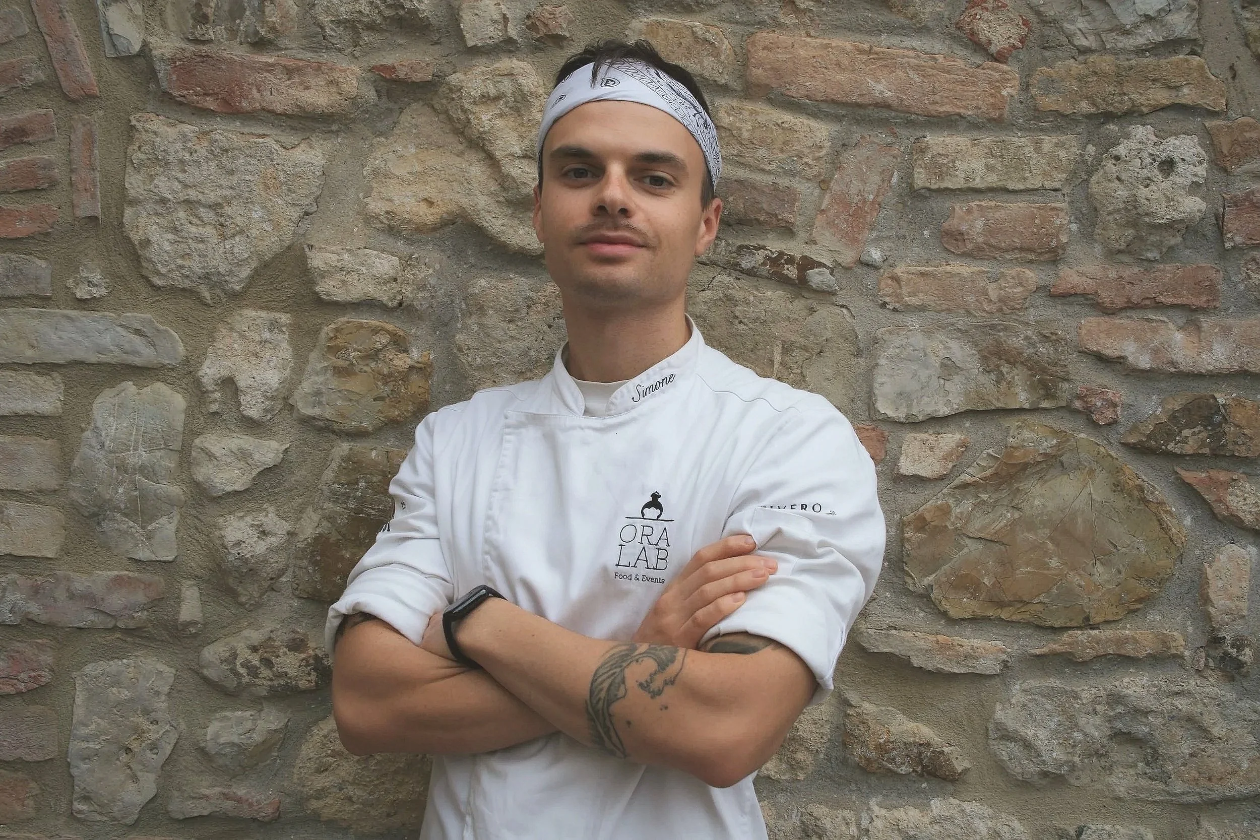 Male chef posing with arms crossed in front of a stone wall, wearing a white chef's jacket with the name 'Simone' embroidered on it, a bandana on his head, and a watch on his left wrist.