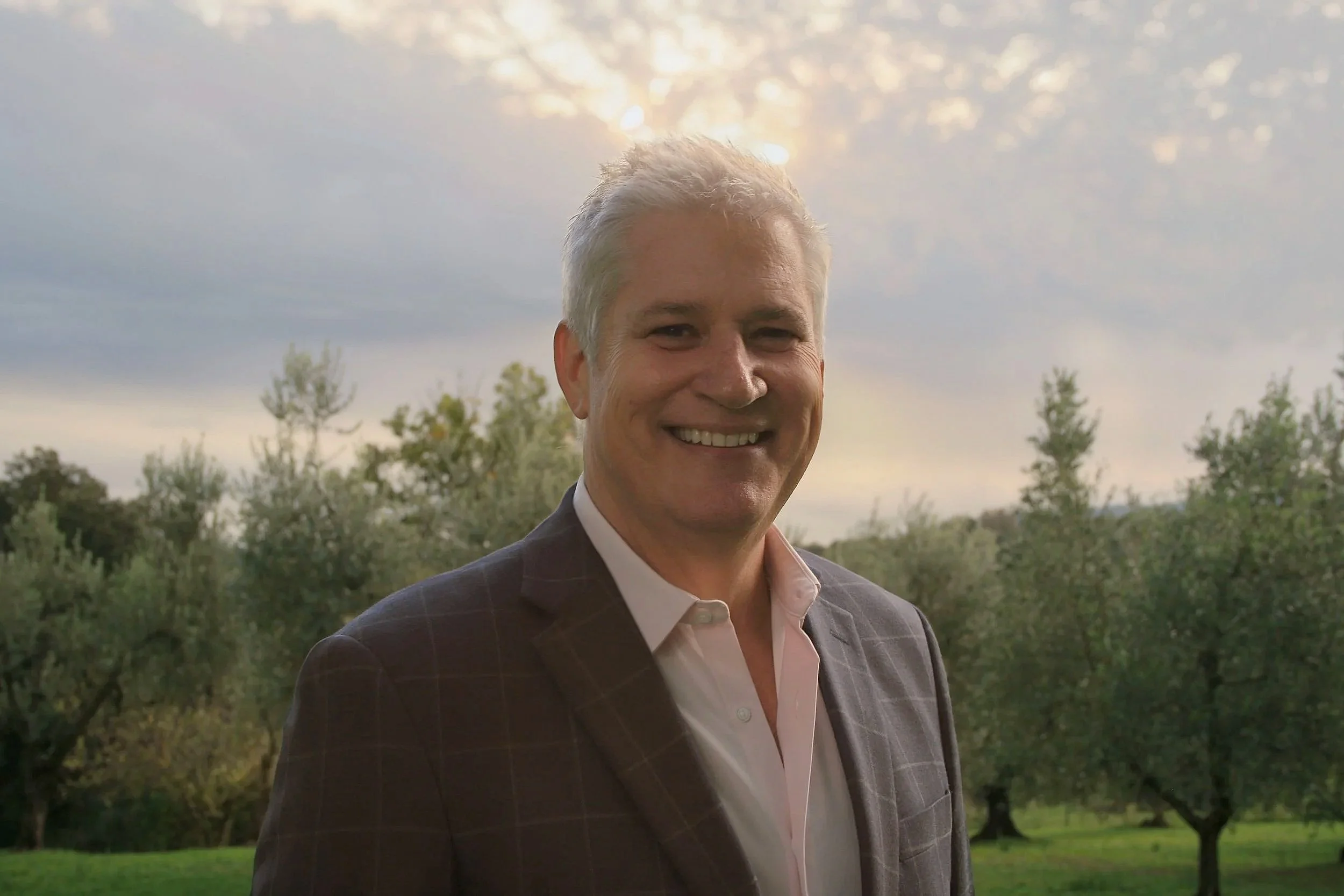 A smiling middle-aged man with gray hair wearing a dark plaid jacket and a white shirt standing outdoors in a park with trees and a cloudy sky in the background.