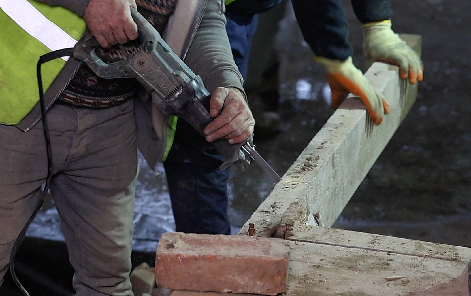 Worker using a power drill to secure a wooden beam, with another worker aligning the beam and wearing gloves, at a construction site.