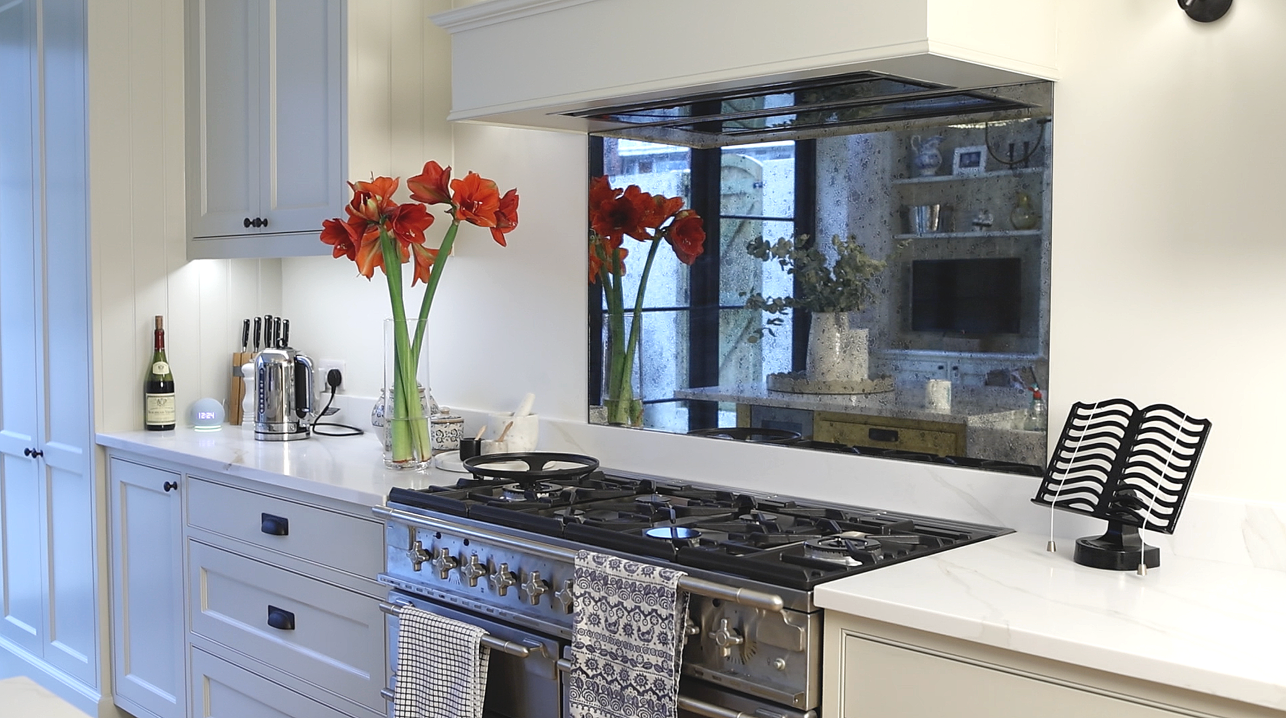 A modern kitchen with white cabinets, a white countertop, and a stainless steel stove. There are two tall vases with red and orange flowers on the countertop. A bottle of wine, a kettle, and a knife block are also visible. A large mirror behind the f