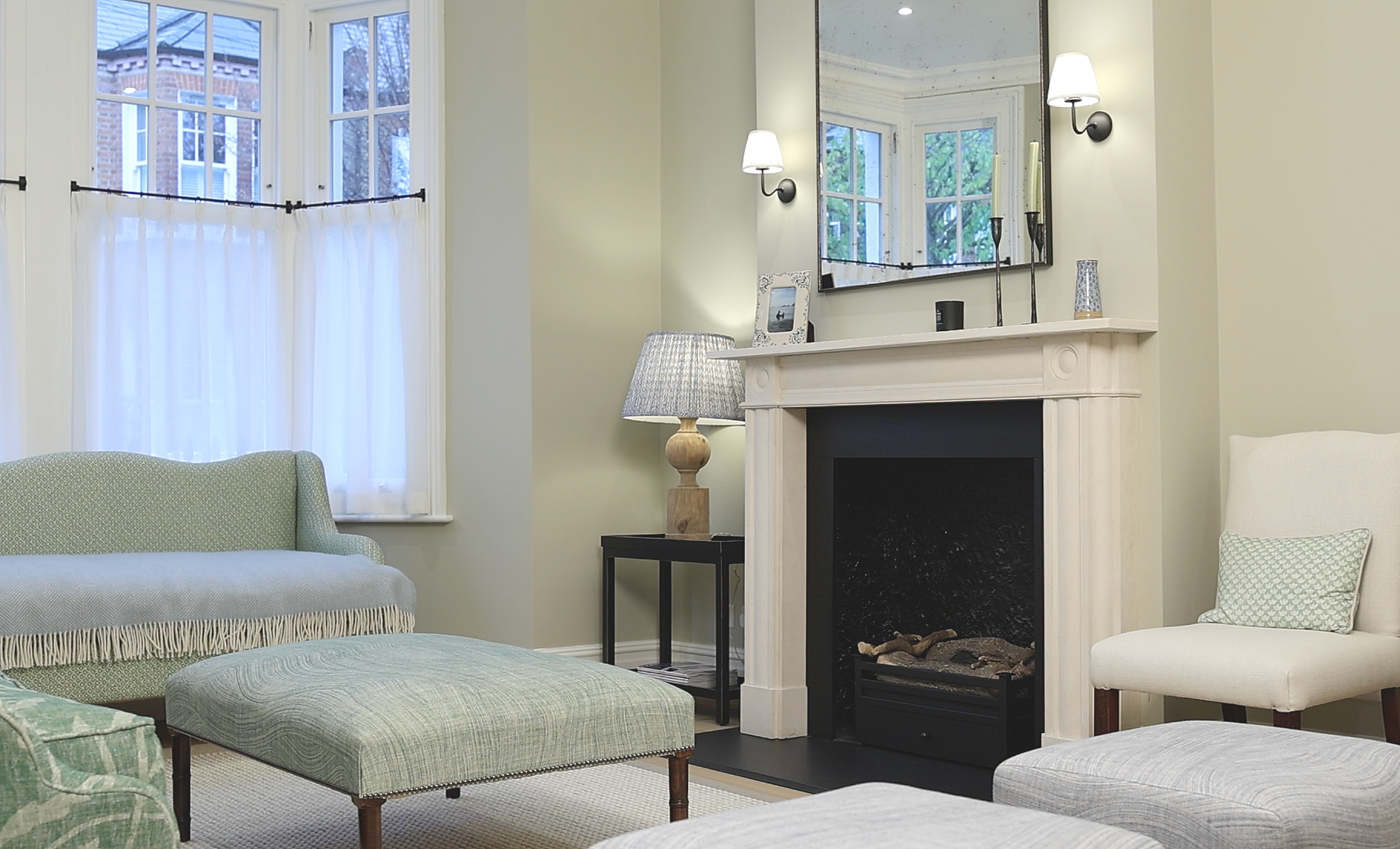 Living room with a fireplace, a mirror above it, a small side table with a lamp, a chair with a pillow, a green upholstered ottoman, and a window with white curtains.
