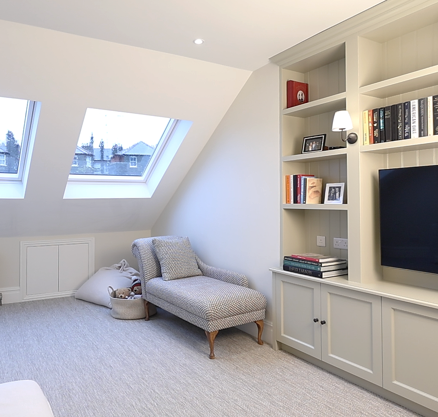 A cozy attic living room with skylight windows, a patterned chaise lounge, a bookshelf with books and framed photos, and a flat-screen TV.