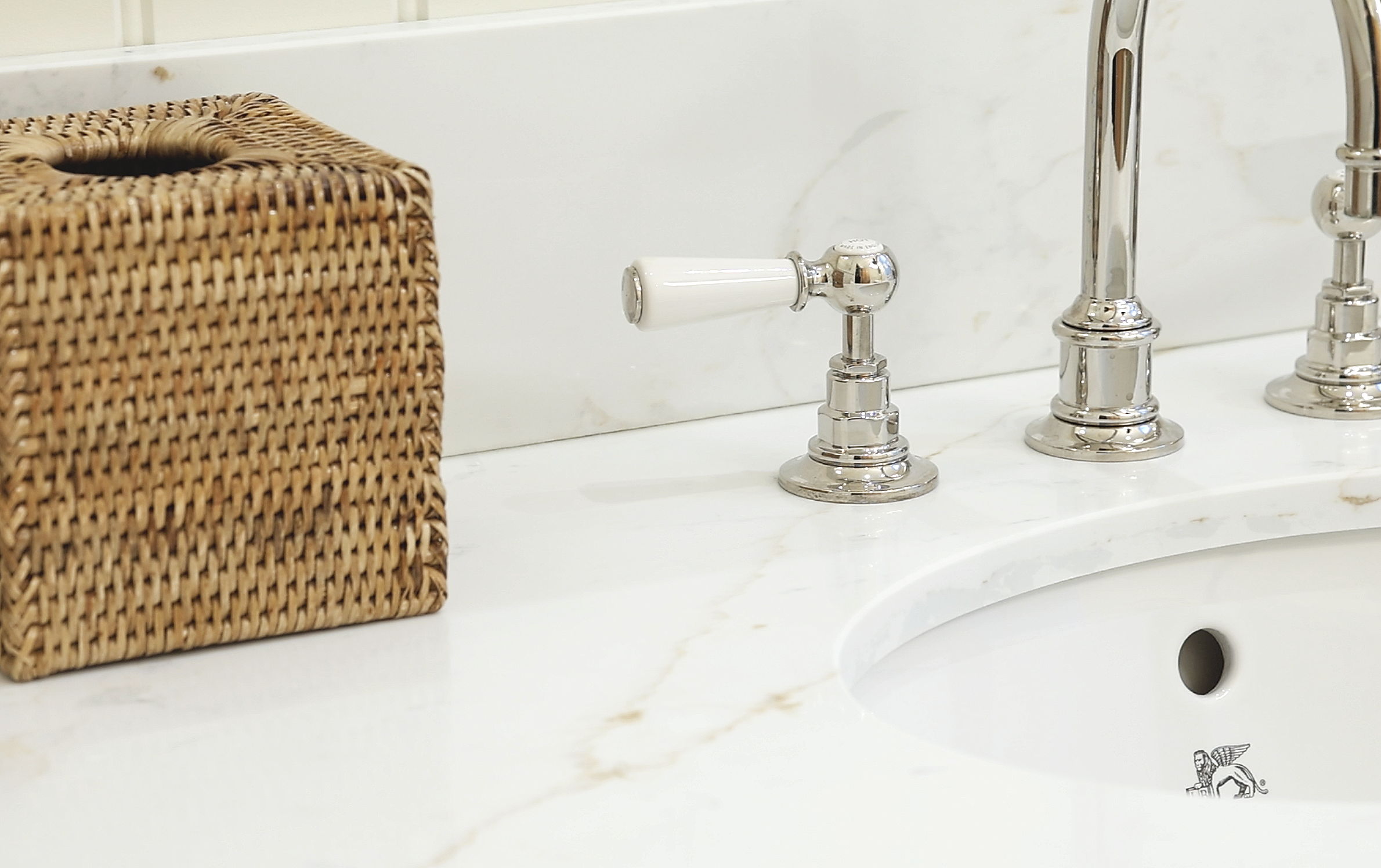 Close-up of a bathroom sink with a woven tissue box on the left, a white ceramic handle faucet, and a chrome faucet on a marble countertop.