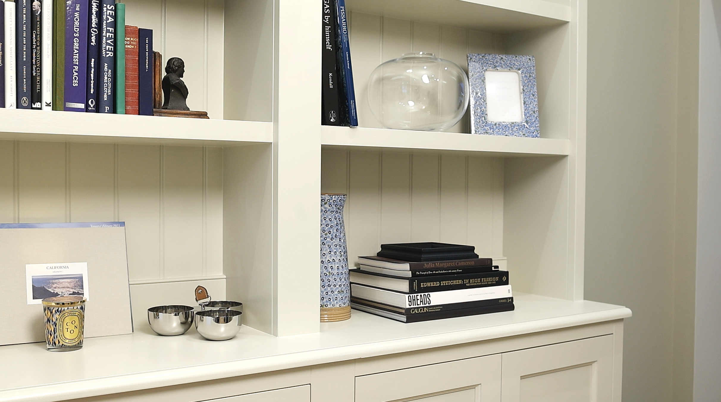 A white bookshelf with books, decorative items including a glass bowl, picture frames, a small bust, and stacked books, some with blue patterned objects and small silver containers on the lower shelf.