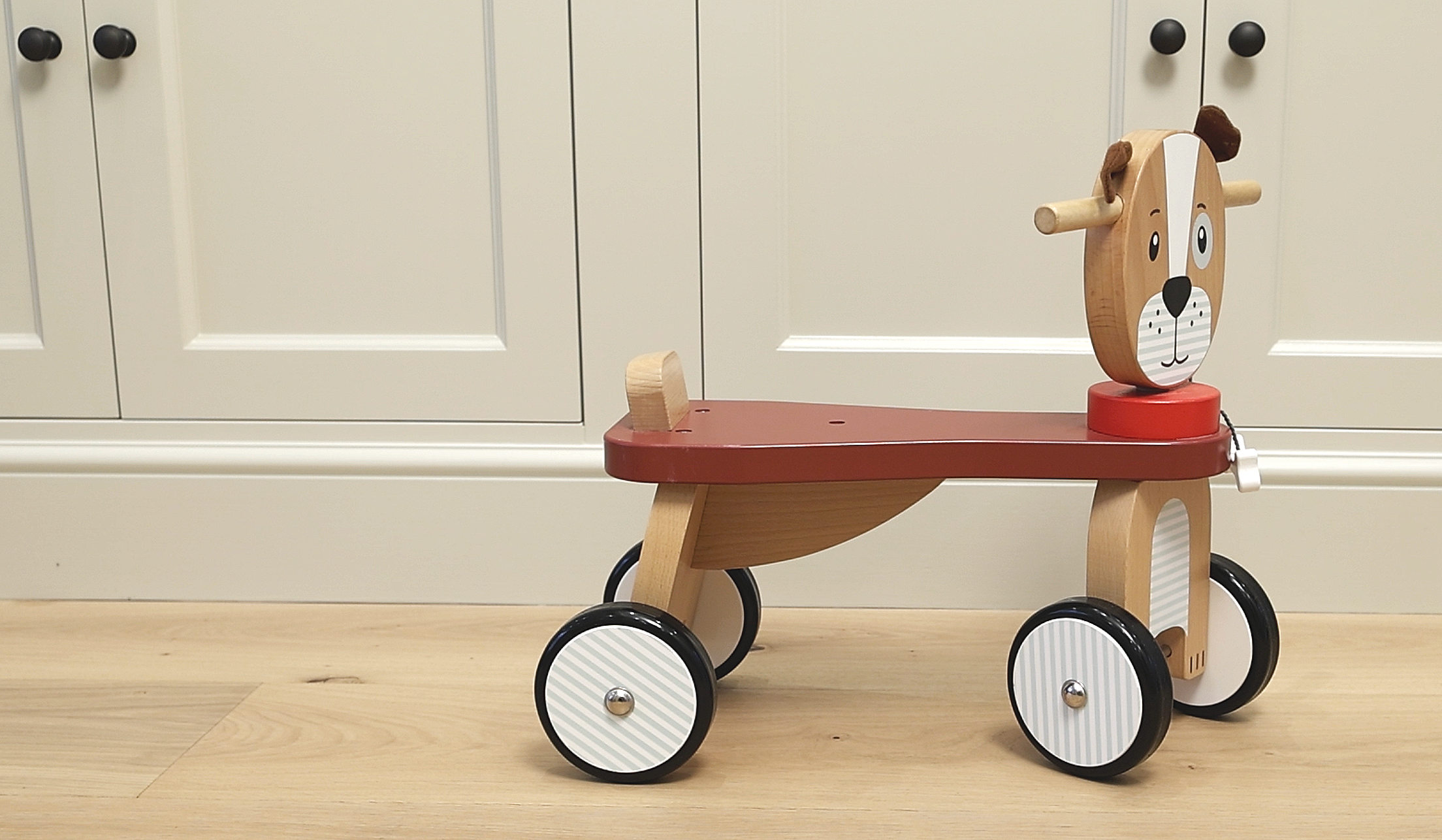 Wooden toy dog on wheels with a red base, striped wheels, and a smiling face, placed on a wooden floor against a white cabinet background.