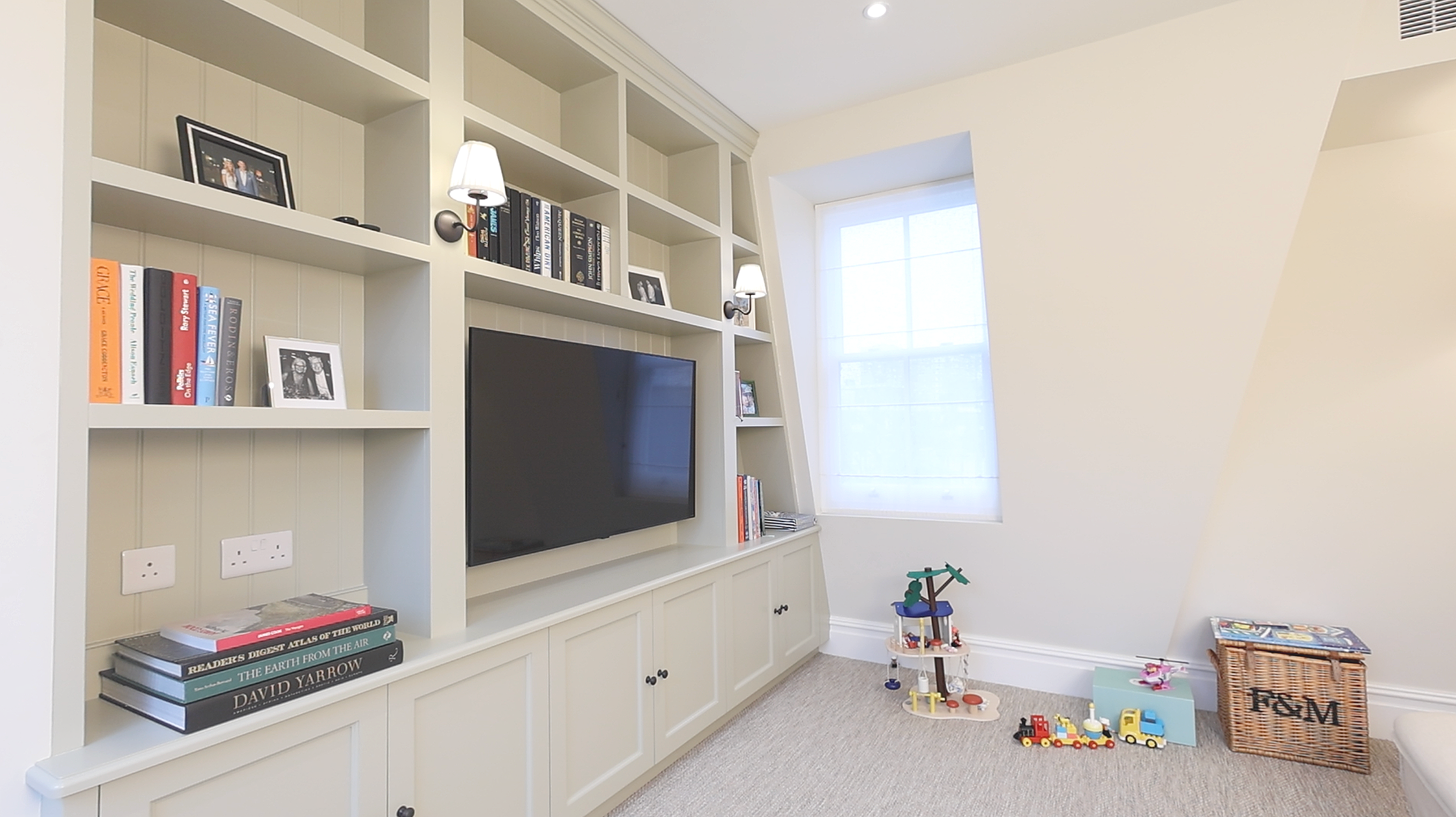 A living room with built-in white shelves and cabinets, a black flat-screen TV, books, photo frames, and small wall lamps. There is a window with sheer white curtains, and children's toys on the beige carpet, including LEGO trains and a playset with 