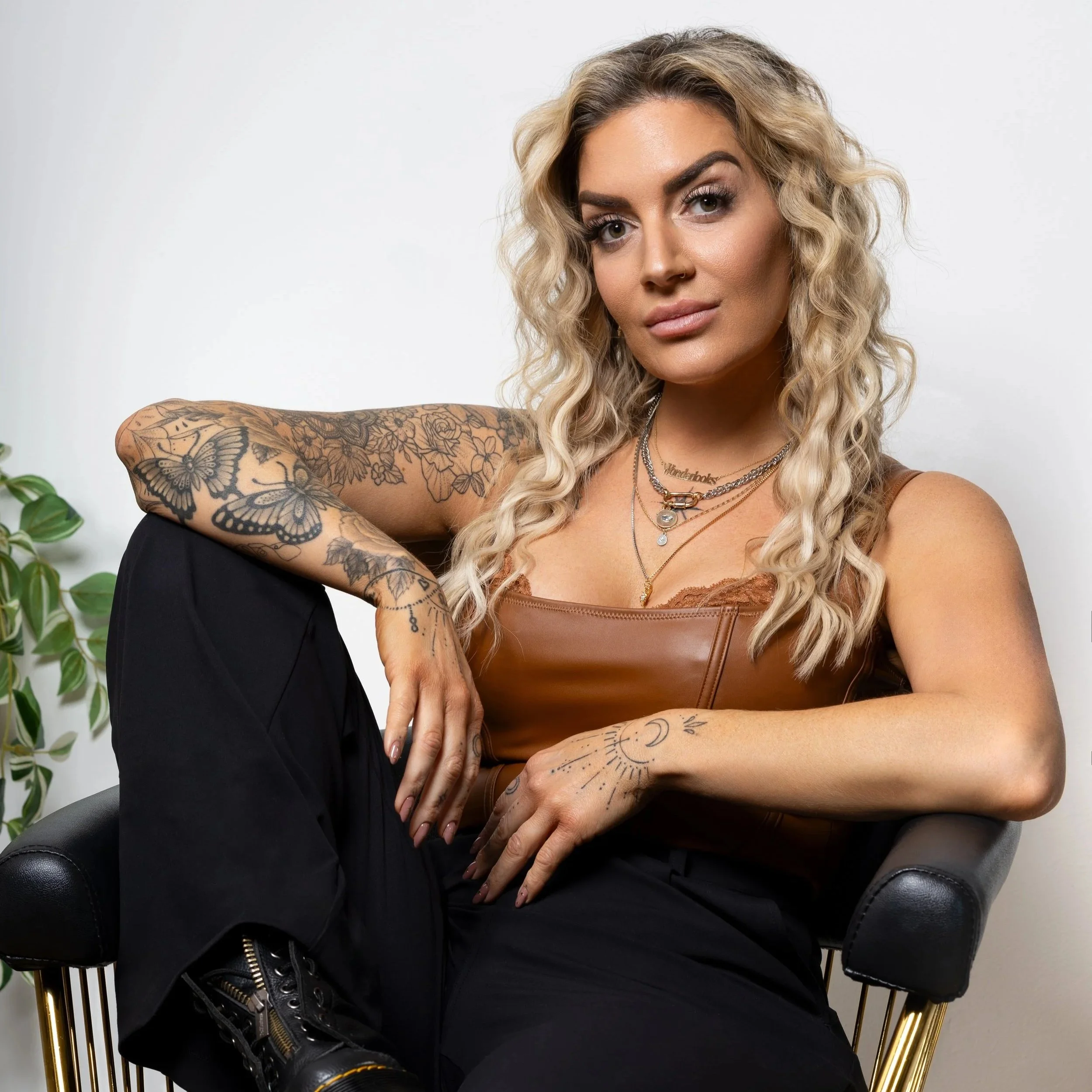 A woman with blonde, curly hair wearing a brown leather top and black pants, sitting on a black chair with gold accents, posing with her arm resting on her knee, showing tattoos on her arm and hand, and wearing layered necklaces.