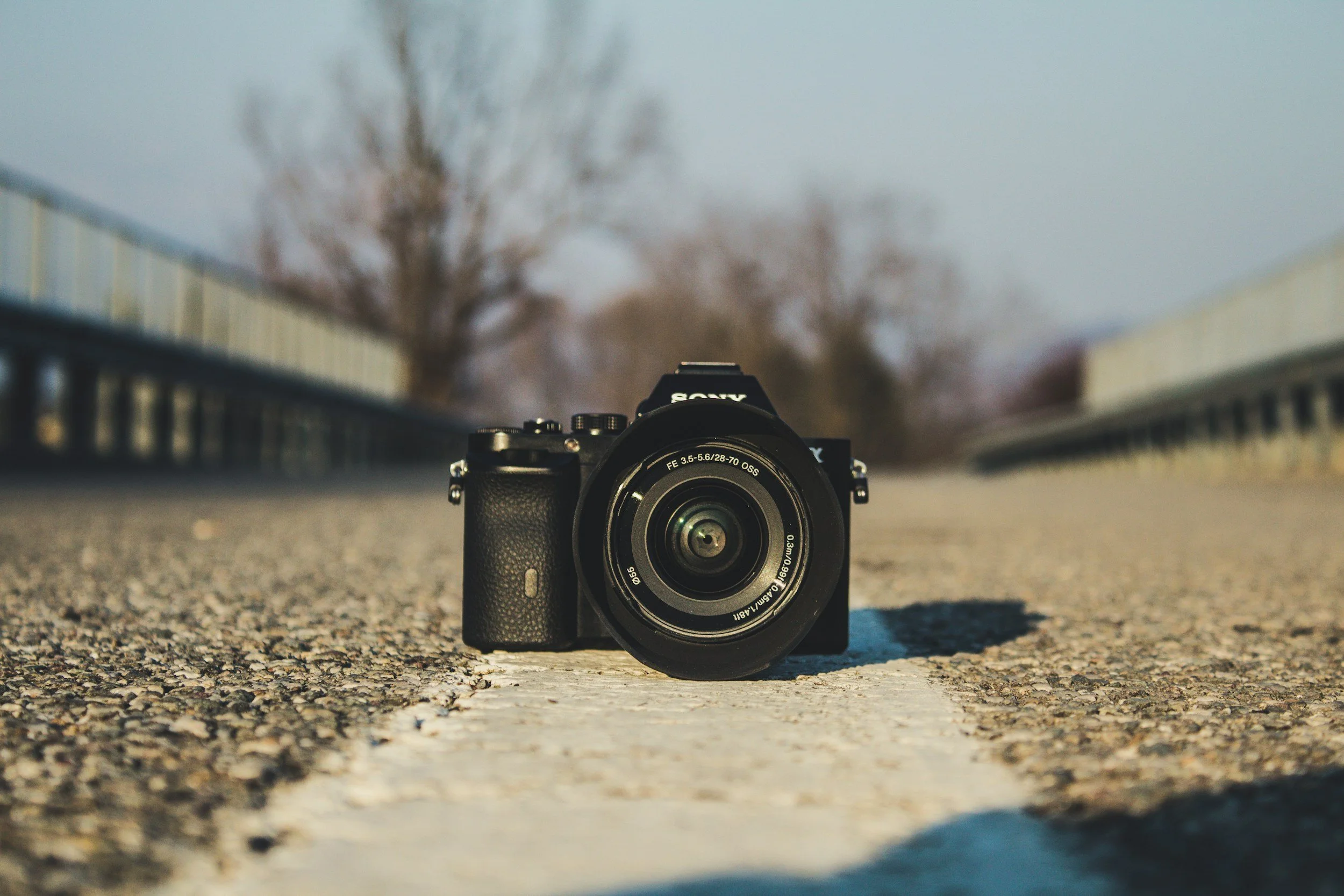 Camera with a black body and lens, placed on a textured roadway with a white line, outdoors during daytime, with blurred trees and sky in the background.