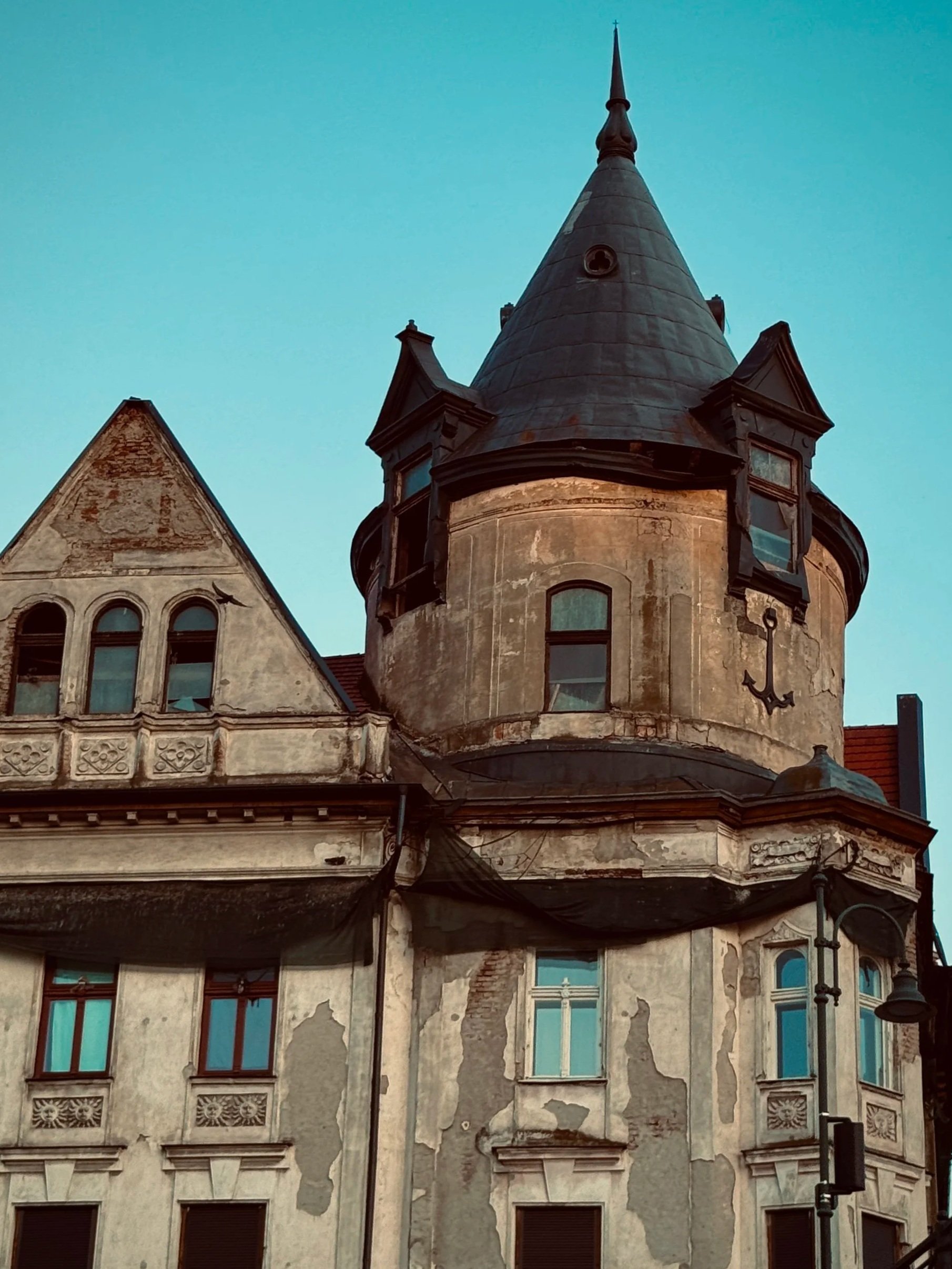 An old building with a round turret and a conical roof, showing peeling paint and weathered walls, with multiple windows and a large anchor symbol on the turret's wall.