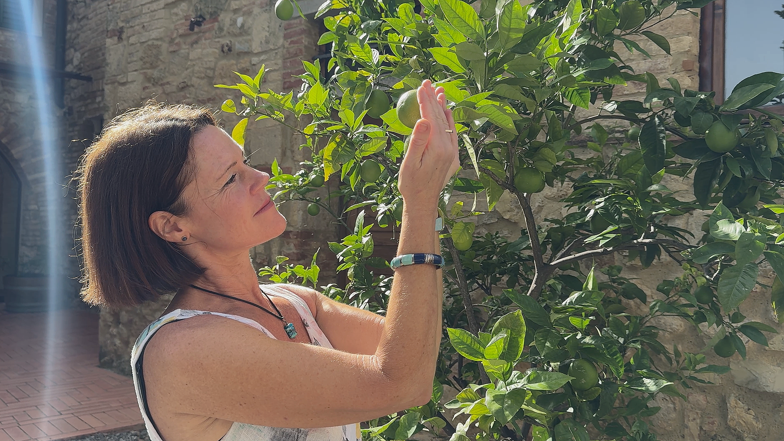 A woman with short brown hair wearing a floral top, a bracelet, and a necklace is standing outdoors, reaching up to a green citrus fruit on a tree with green leaves, against a background of a stone wall and a window.