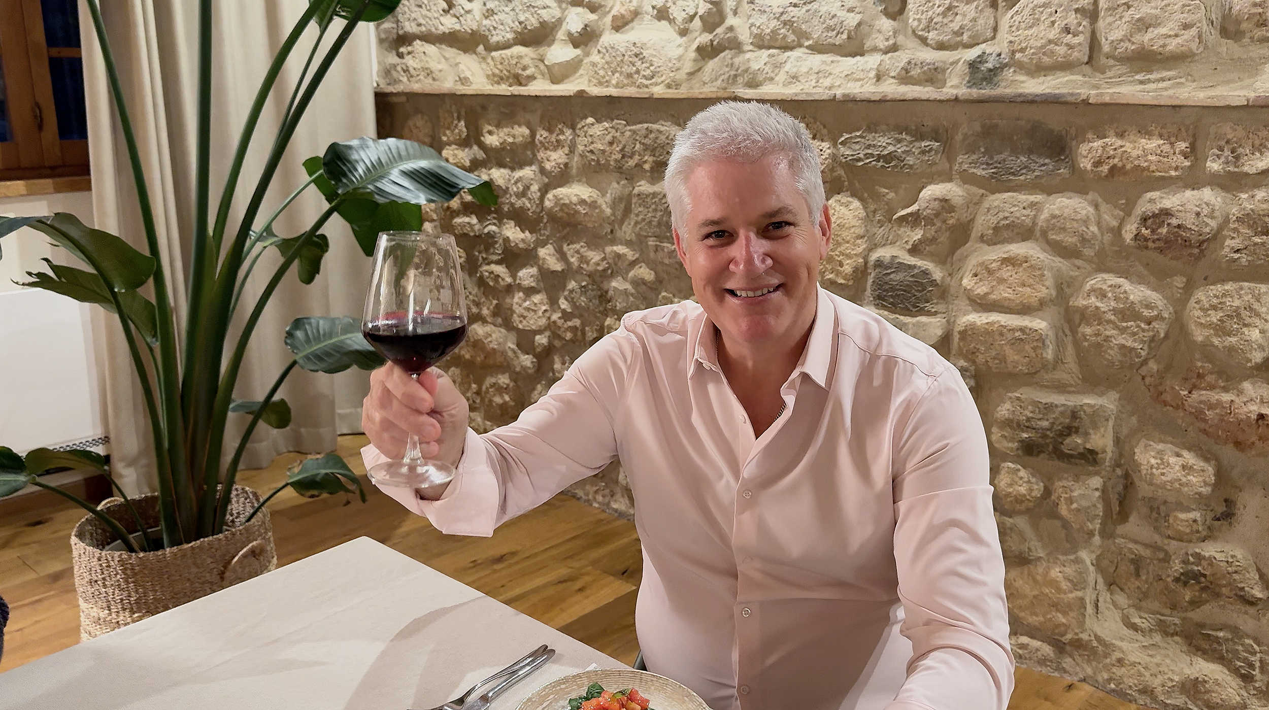 A smiling man with short gray hair in a light pink shirt holding a glass of red wine while sitting at a dining table. There is a large green plant with broad leaves in a woven pot on the left side of the image. The background features a stone wall, a