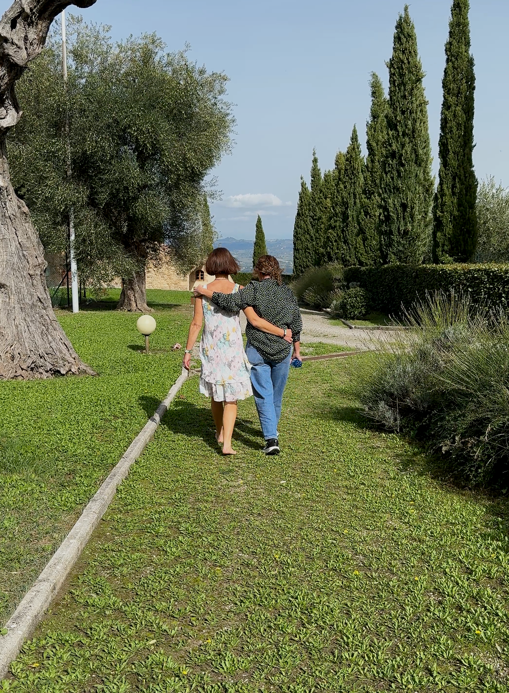 Two women walk together on a lush green path lined with tall cypress trees and shrubs, surrounded by large trees and modern landscape lighting under a clear blue sky.
