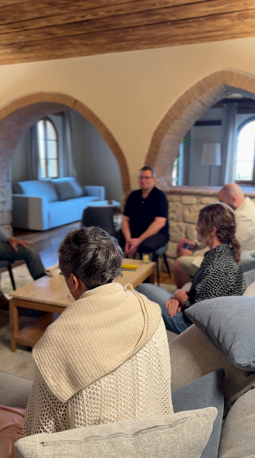 Group of people sitting in a cozy living room with exposed brick arches, hardwood floors, and large windows, having a discussion.