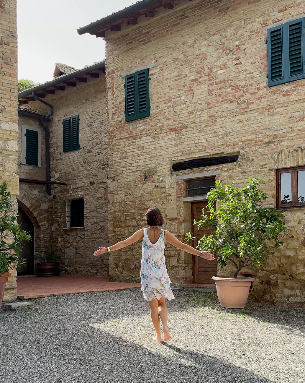 A woman with shoulder-length dark hair in a floral summer dress walking barefoot on a gravel courtyard, arms outstretched, next to large potted trees in front of an old stone building with green shutters.