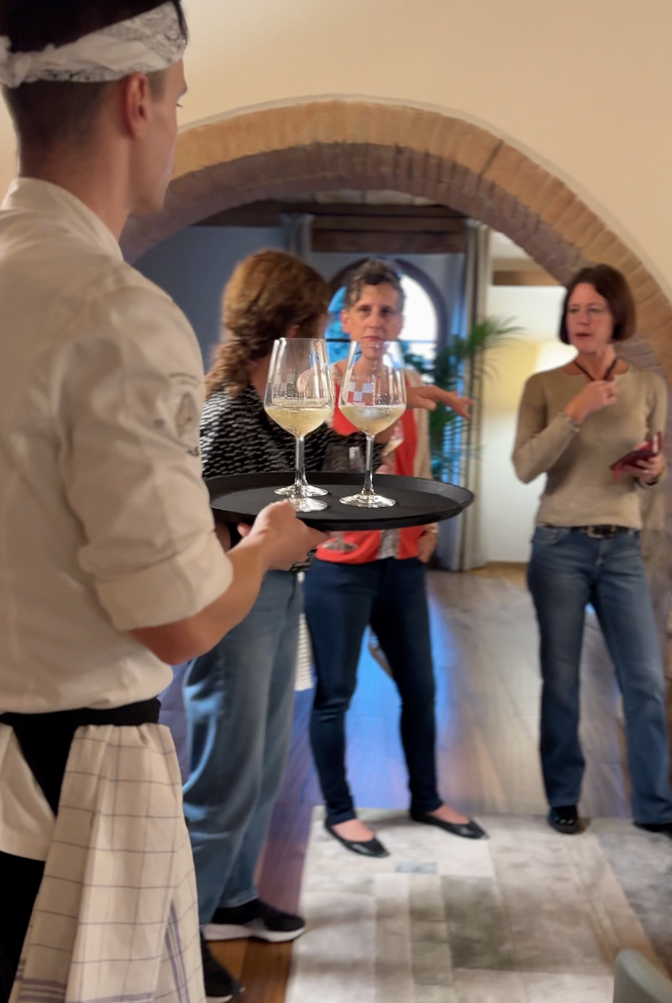A waiter holding a tray with two glasses of white wine in front of three women in conversation inside a restaurant or cafe with arched brick ceiling.