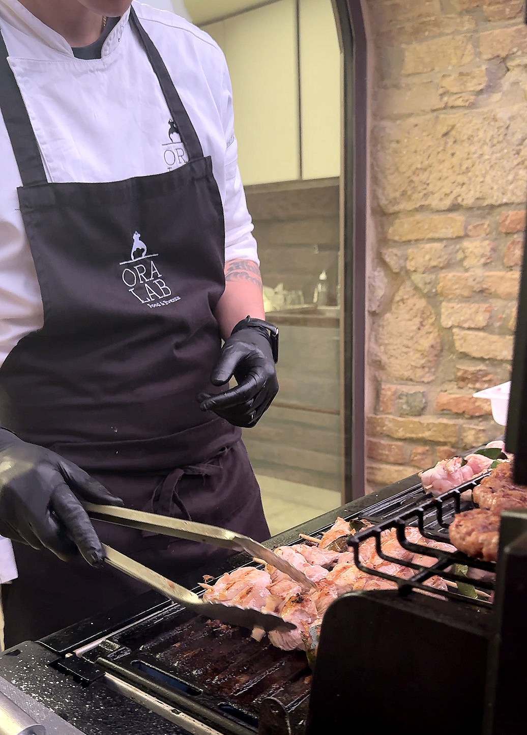 Person wearing a white chef’s coat, black apron with 'ORA LAB' logo, and black gloves, grilling meat on a barbecue grill.