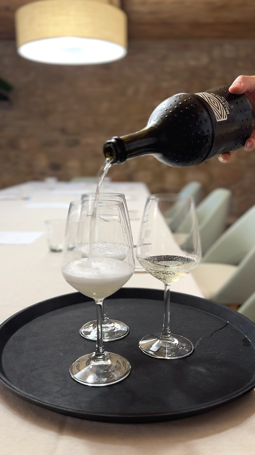 A person pouring white wine from a dark bottle into two wine glasses on a round black tray. The setting appears to be a dining room with chairs and a brick wall in the background.