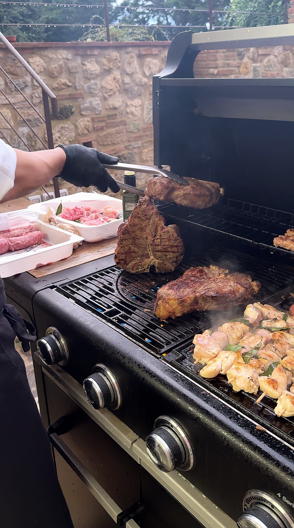 Person wearing black gloves grilling large cuts of beef and chicken skewers on a barbecue grill, with raw meats on the side.