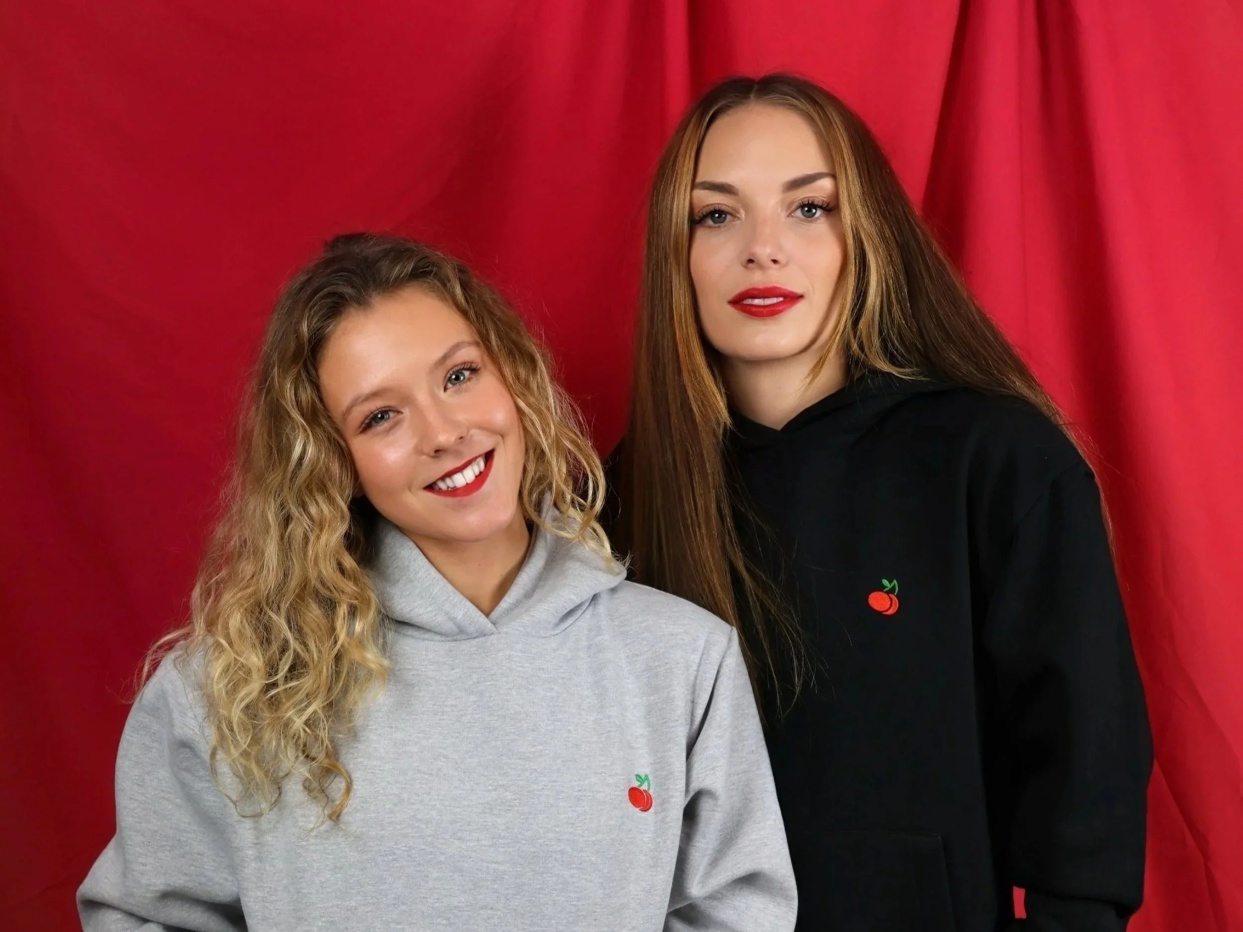 Two young women with light skin, long wavy hair, red lipstick, and casual hoodies, standing in front of a red backdrop.