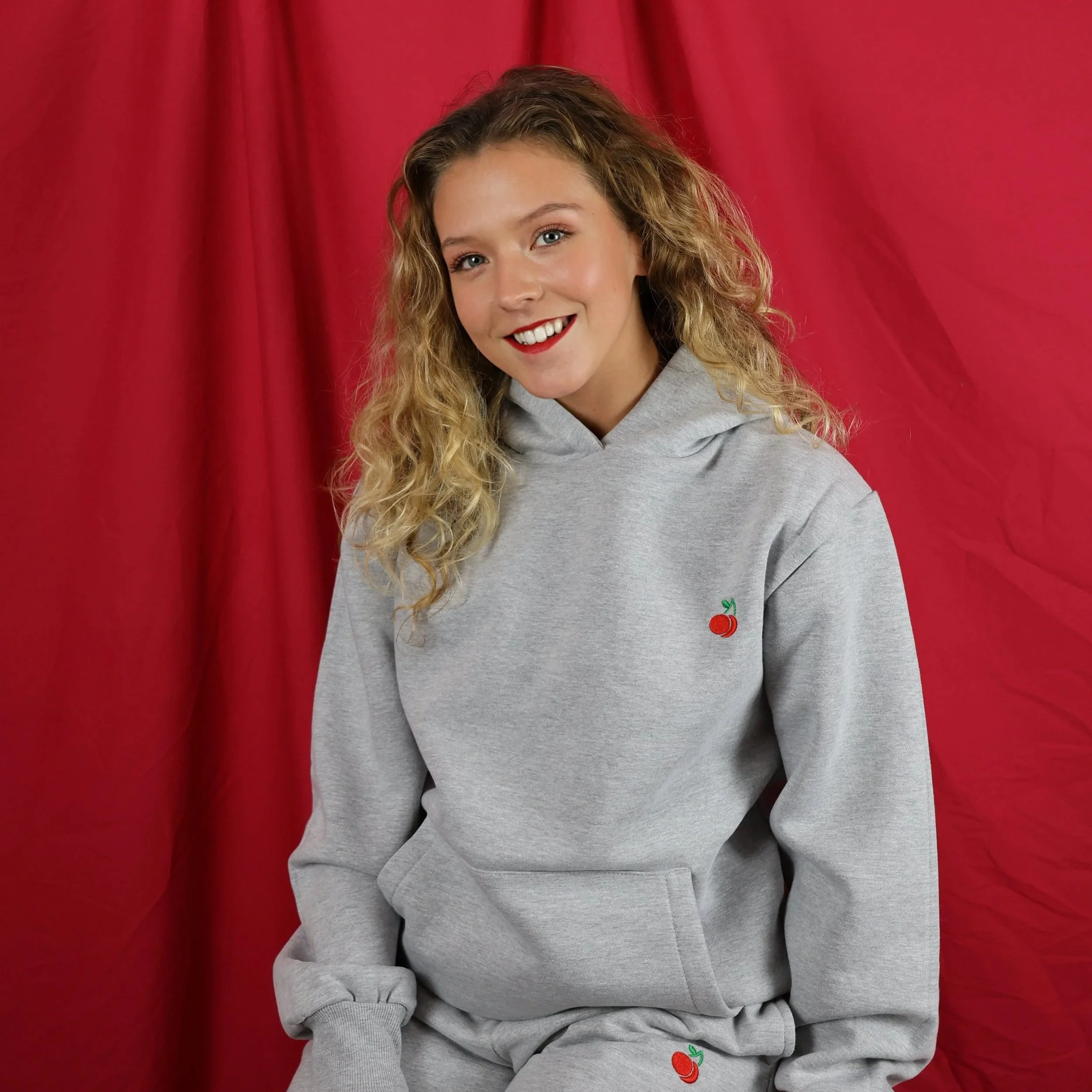 A young woman with curly blonde hair, blue eyes, and red lipstick, smiling, wearing a gray hoodie with a cherry embroidered on it, standing in front of red curtains.