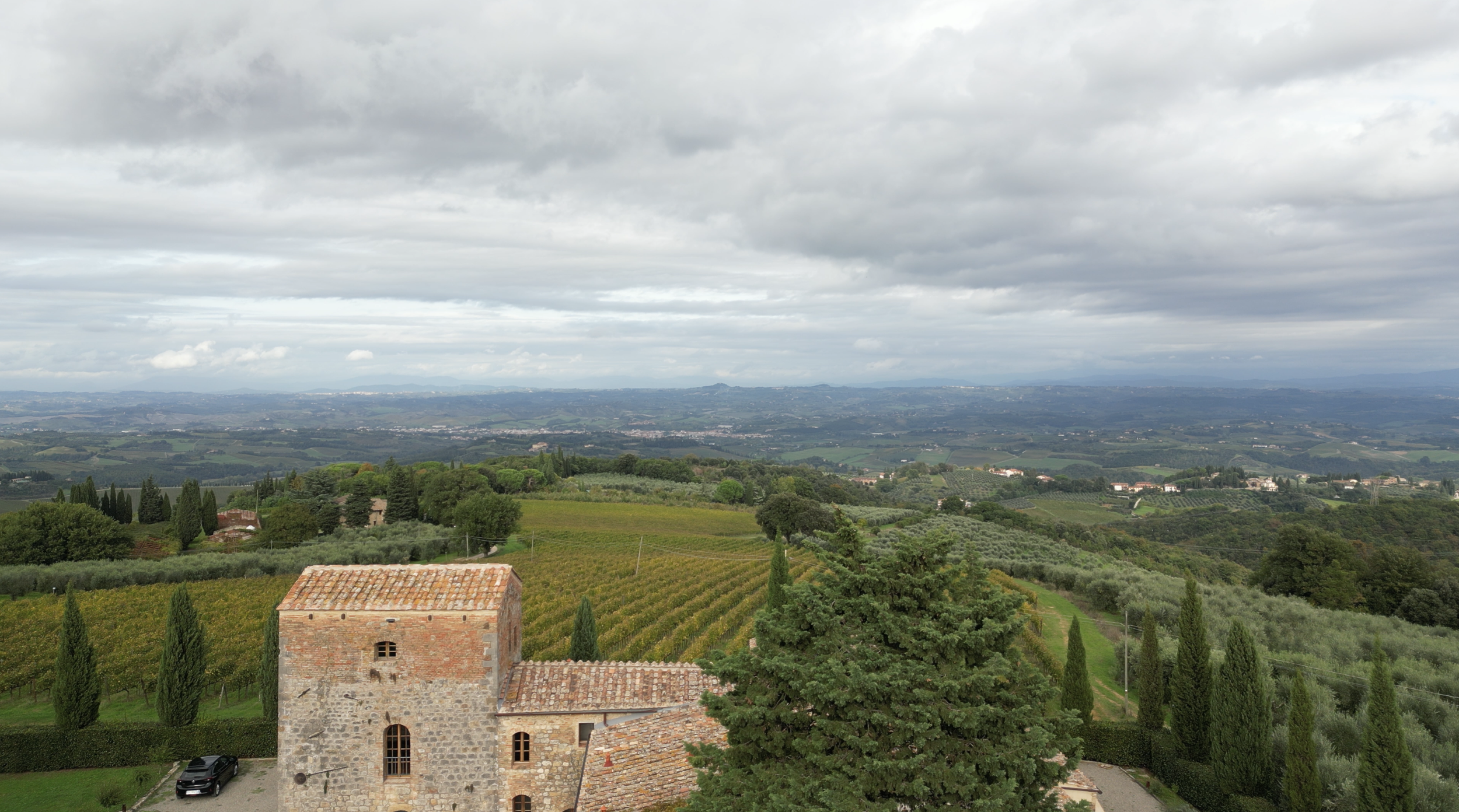 Scenic view of rolling hills with vineyards, trees, and an old stone building, under a cloudy sky in the countryside.