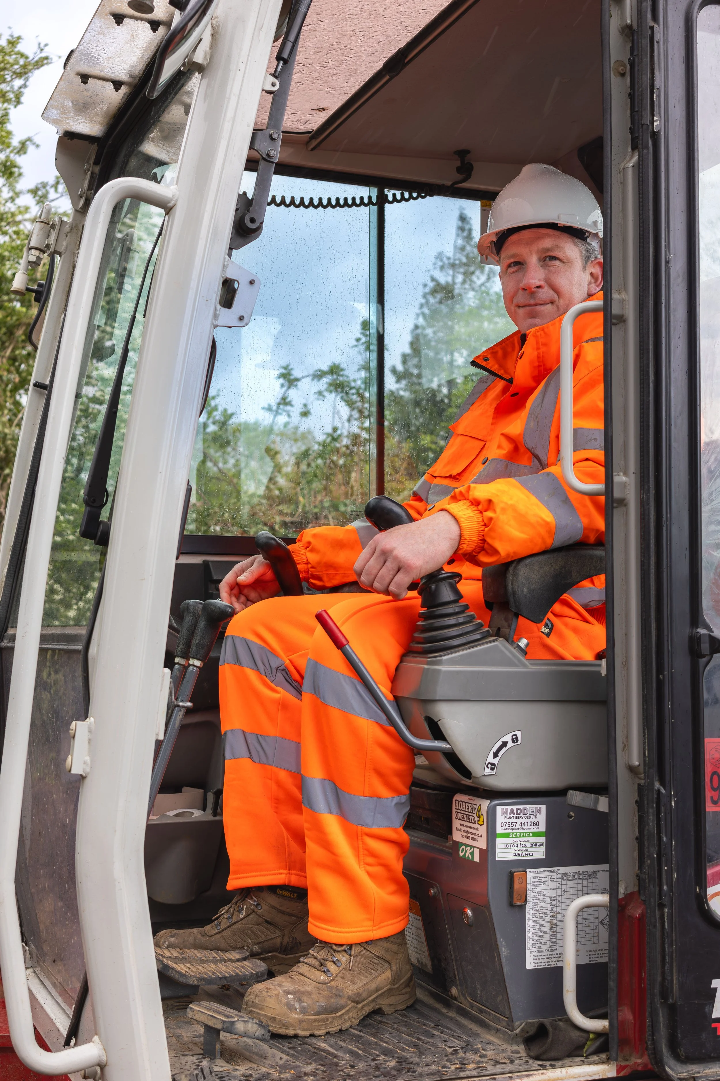 Construction worker operating a skid steer loader, wearing orange safety uniform, white hard hat, and brown work boots, inside the vehicle cab.