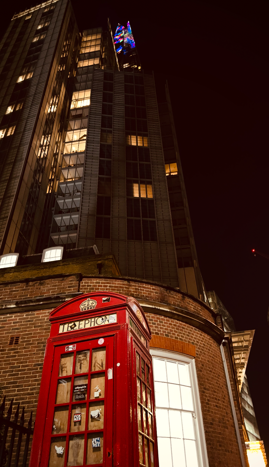 A traditional red telephone booth in front of a modern skyscraper at night with windows illuminated.
