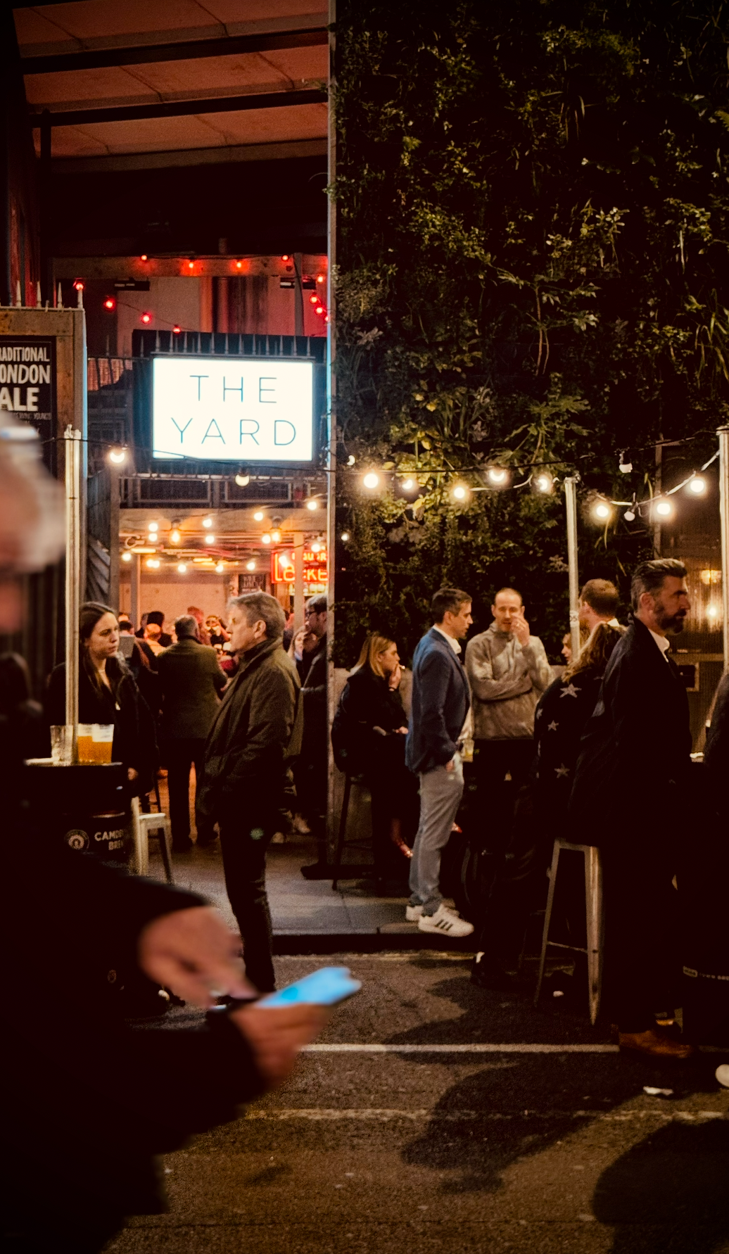 People gathering outdoors at night near a bar or restaurant called The Yard, illuminated by string lights, with some standing and chatting, and the interior visible with more people and warm lighting.