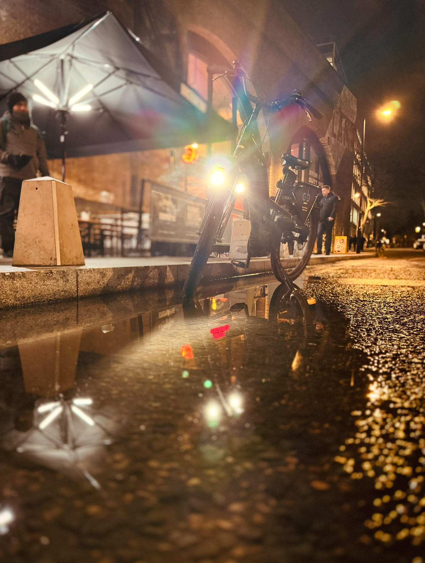 A black bicycle parked on a street with a puddle reflecting the bike's lights and the umbrella behind it. Street lights and a building with brick walls are visible in the background at night.