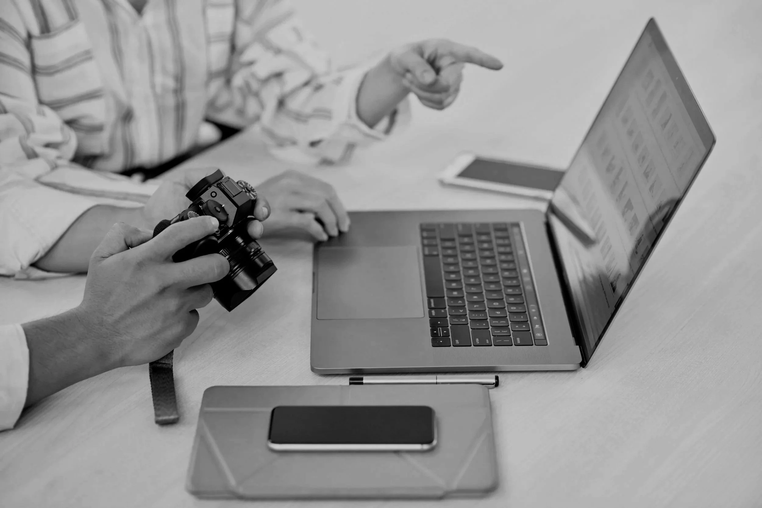 Two people at a desk looking at a laptop screen with one person holding a camera and the other pointing at the screen. Various electronic devices are on the desk.