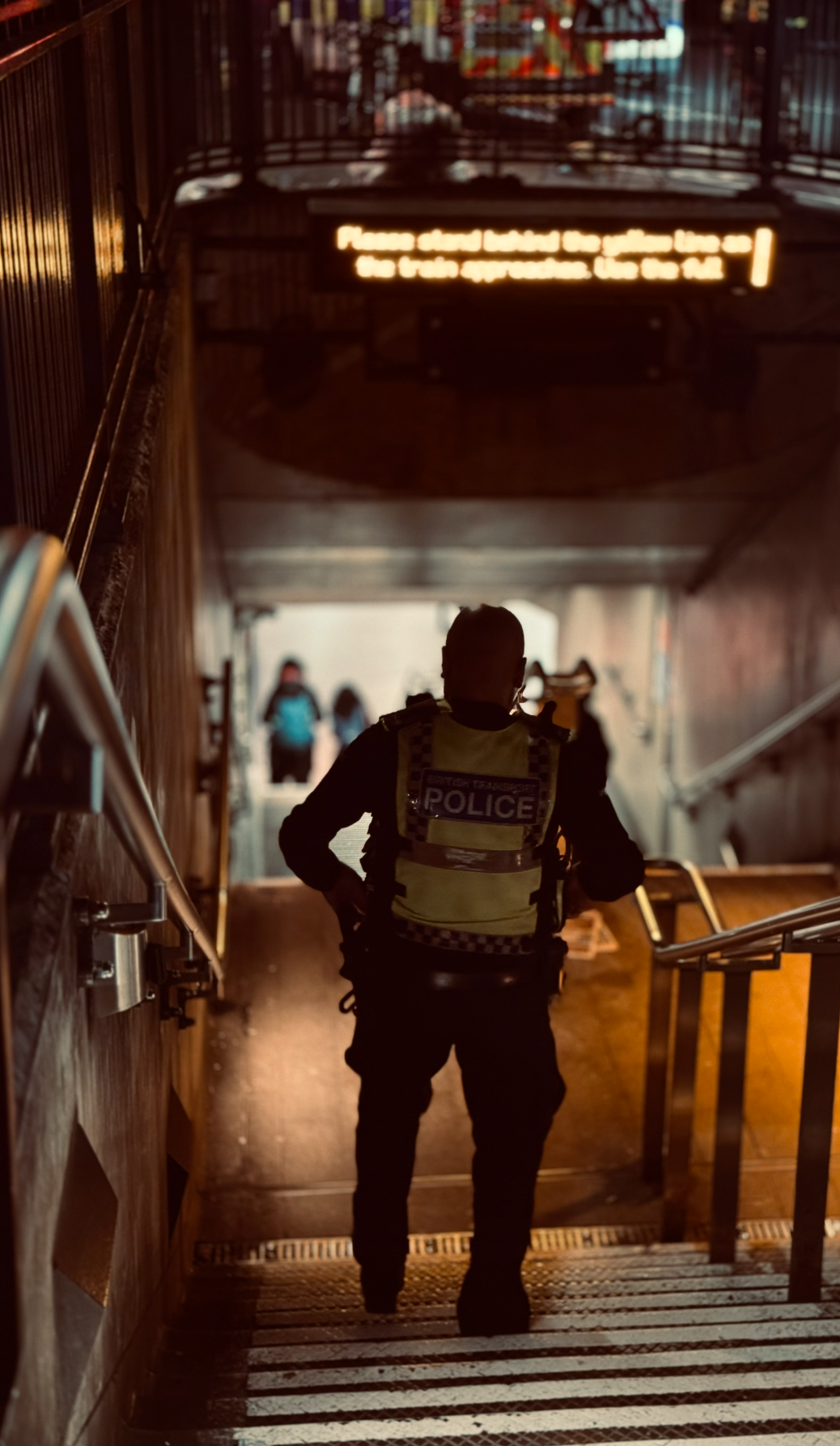A police officer standing at the bottom of a stairway in a public space, with people walking in the background and an illuminated sign overhead.