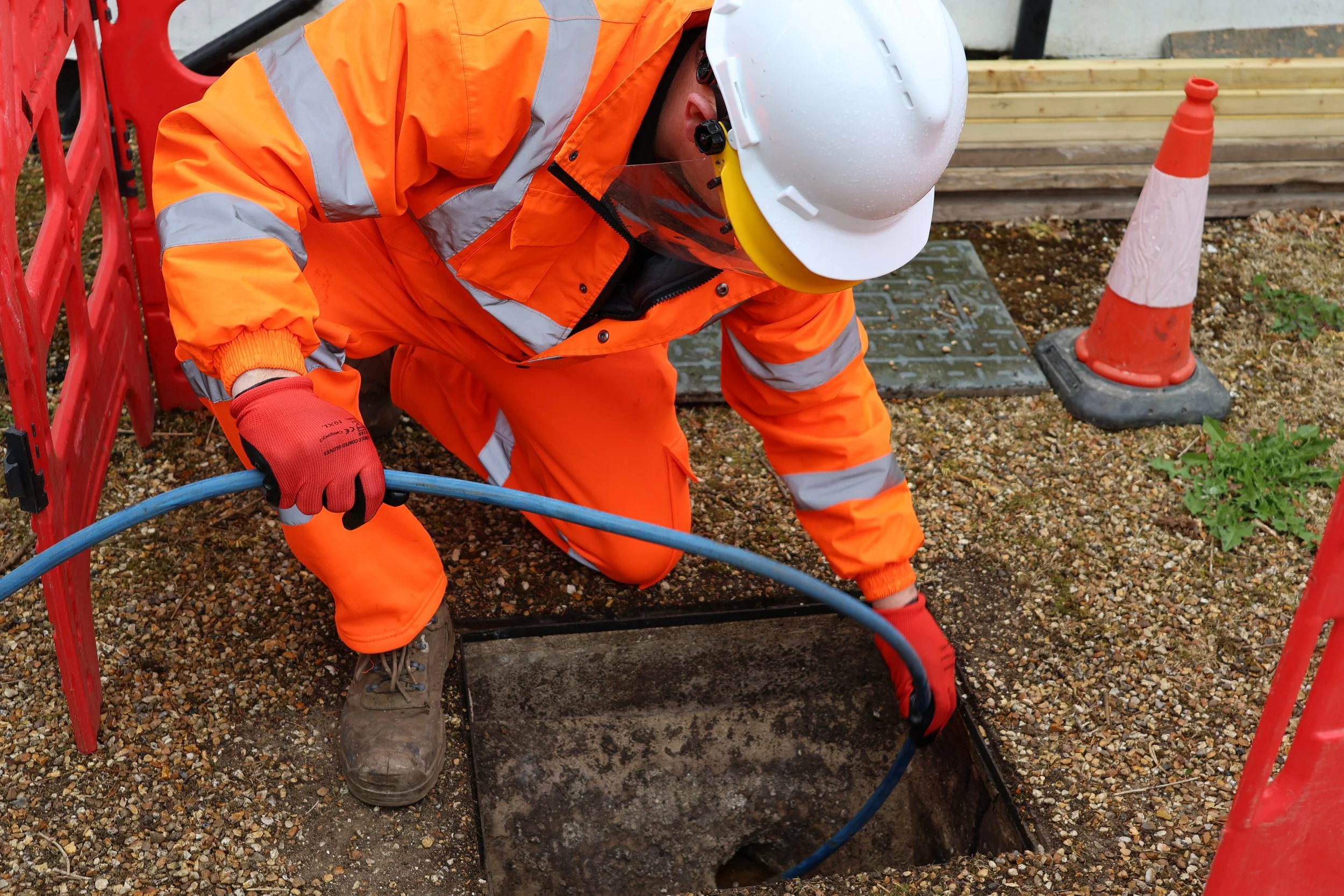 A construction worker wearing an orange safety suit, white hard hat, and red gloves, kneeling and working in an open hole on a construction site, with orange traffic cones and wooden planks nearby.