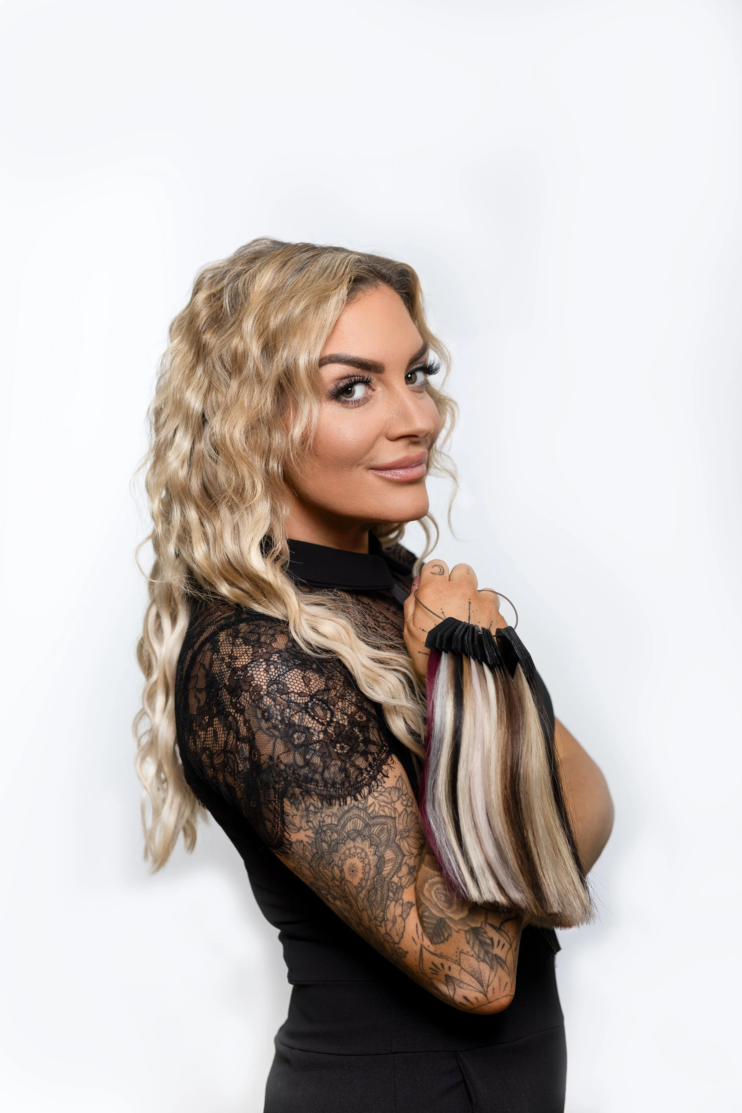 Woman with blonde, curly hair holding a bundle of blonde, black, and purple hair extensions against a white background.