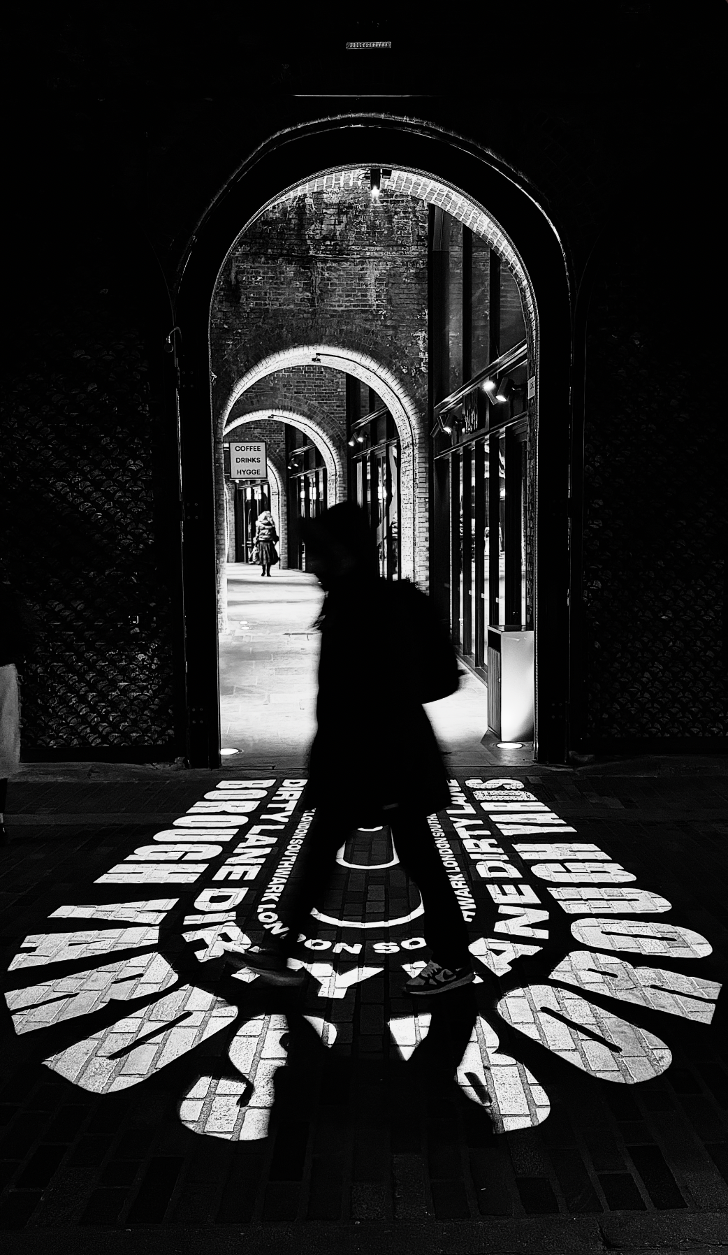 A black and white photo of a person walking through a doorway with light projected onto the ground, displaying the words 'SAFE TOWN, OUR TOWN, HERE TO STAY,' and a spoon icon. The scene is inside a brick building with arched doorways and multiple peo