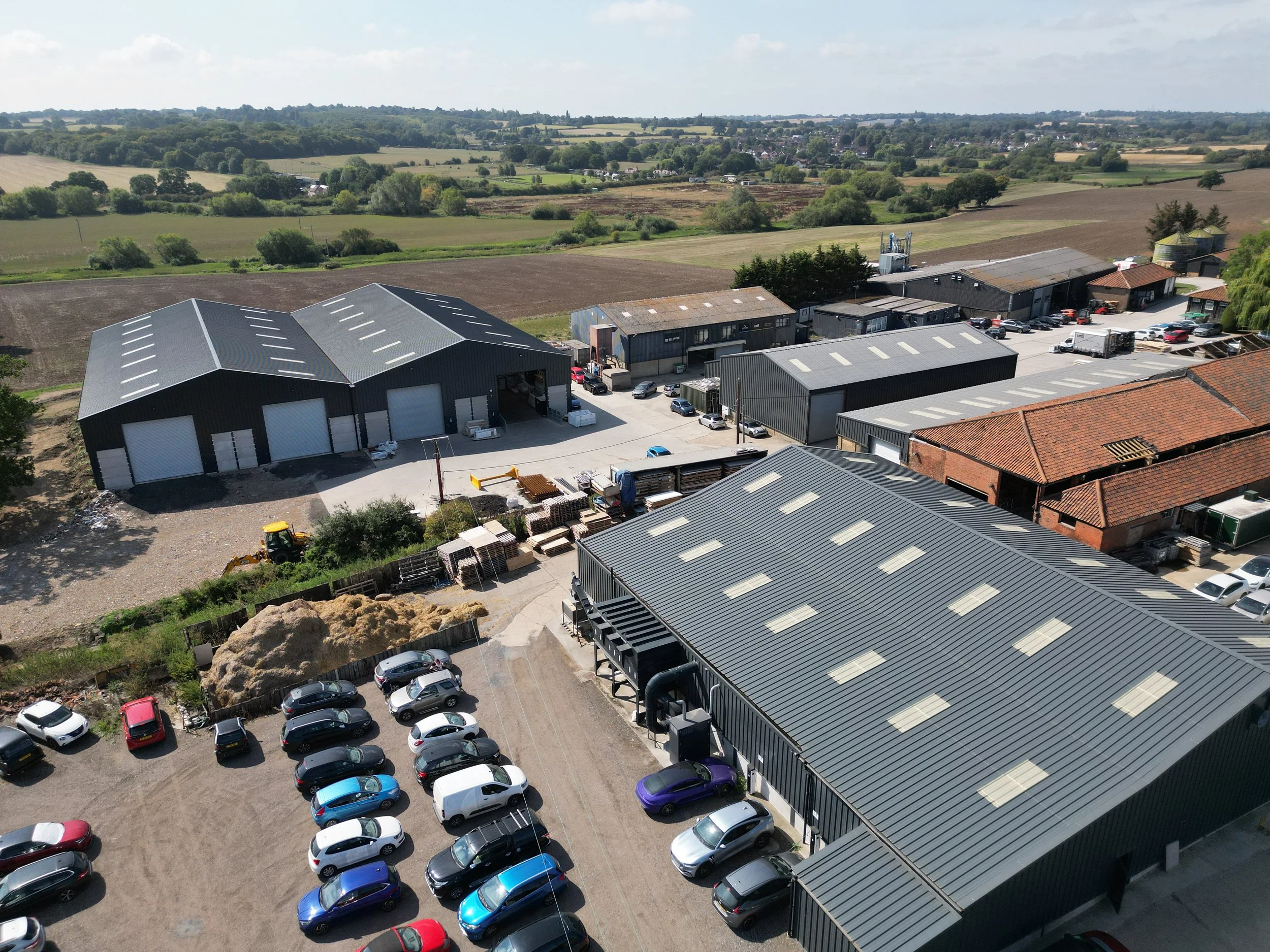 Aerial view of warehouses and buildings in a rural area with parking lots, trees, fields, and open farmland in the background.