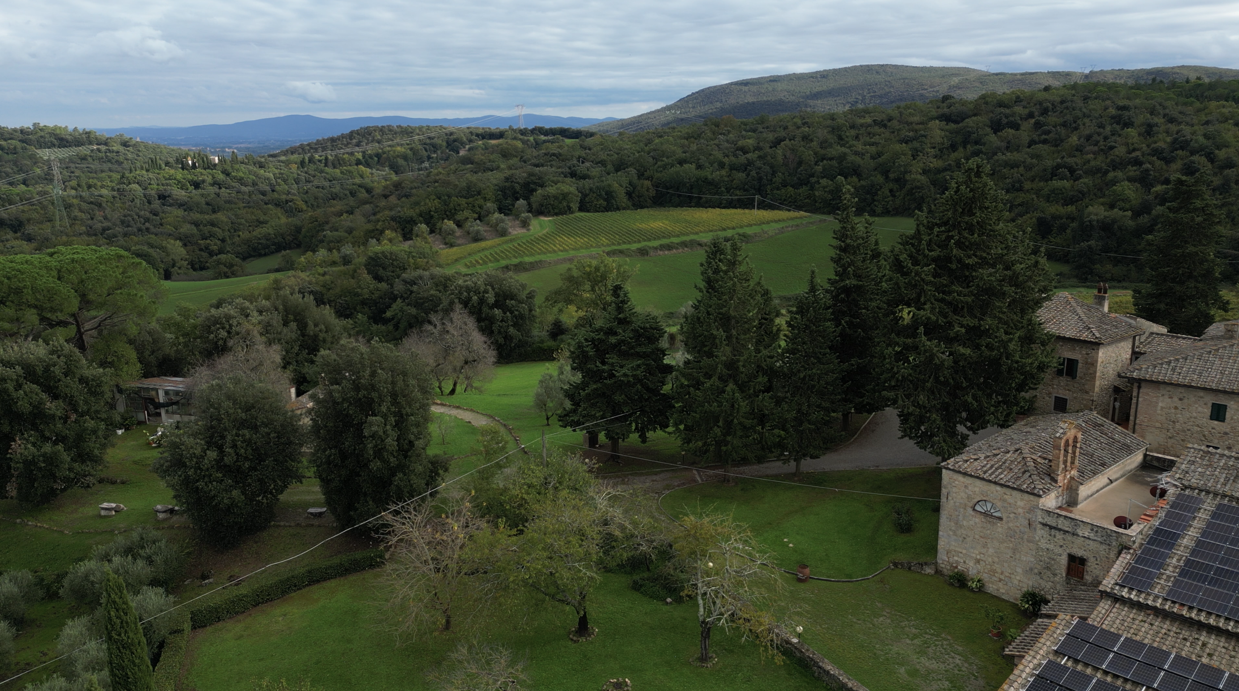A scenic landscape with green rolling hills, trees, and a cluster of old stone buildings with tiled roofs, some of which have solar panels. The background features distant mountains under a cloudy sky.