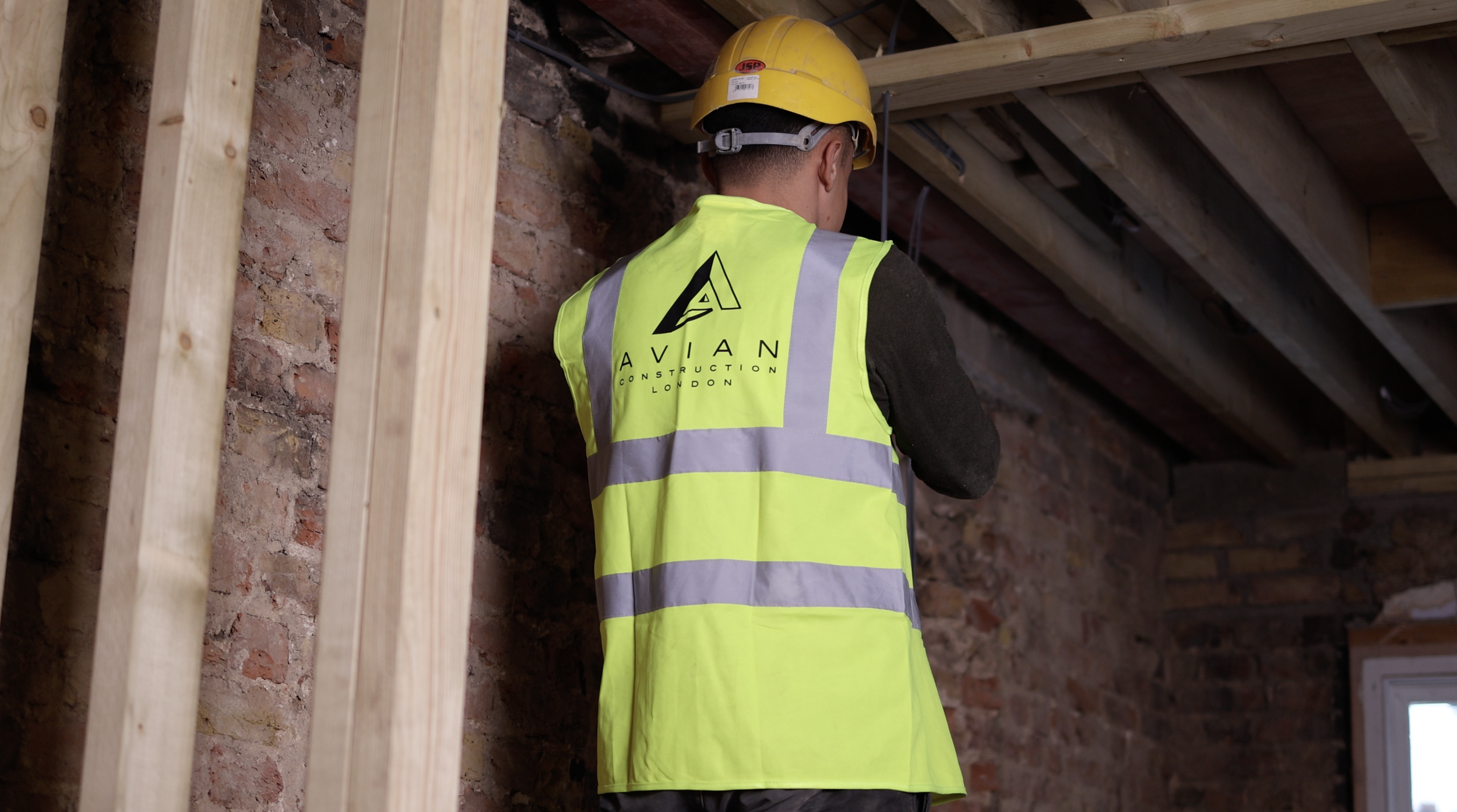 Construction worker wearing a yellow safety helmet and a high-visibility vest with "AVIAN CONSTRUCTION LONDON" logo, working indoors with exposed wooden beams and brick walls.