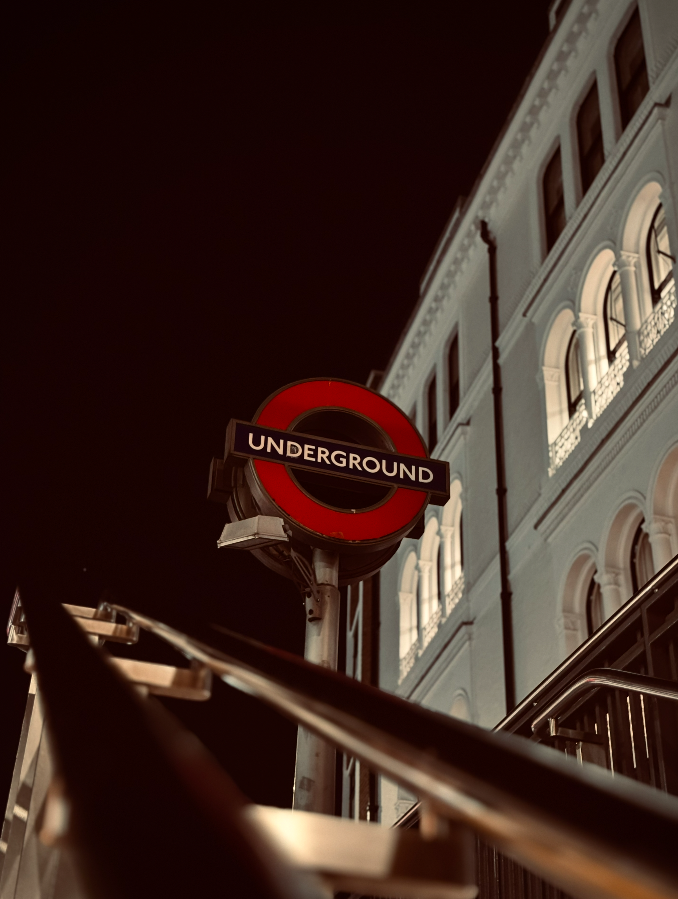 A London Underground sign illuminated at night outside a historic building with arched windows.