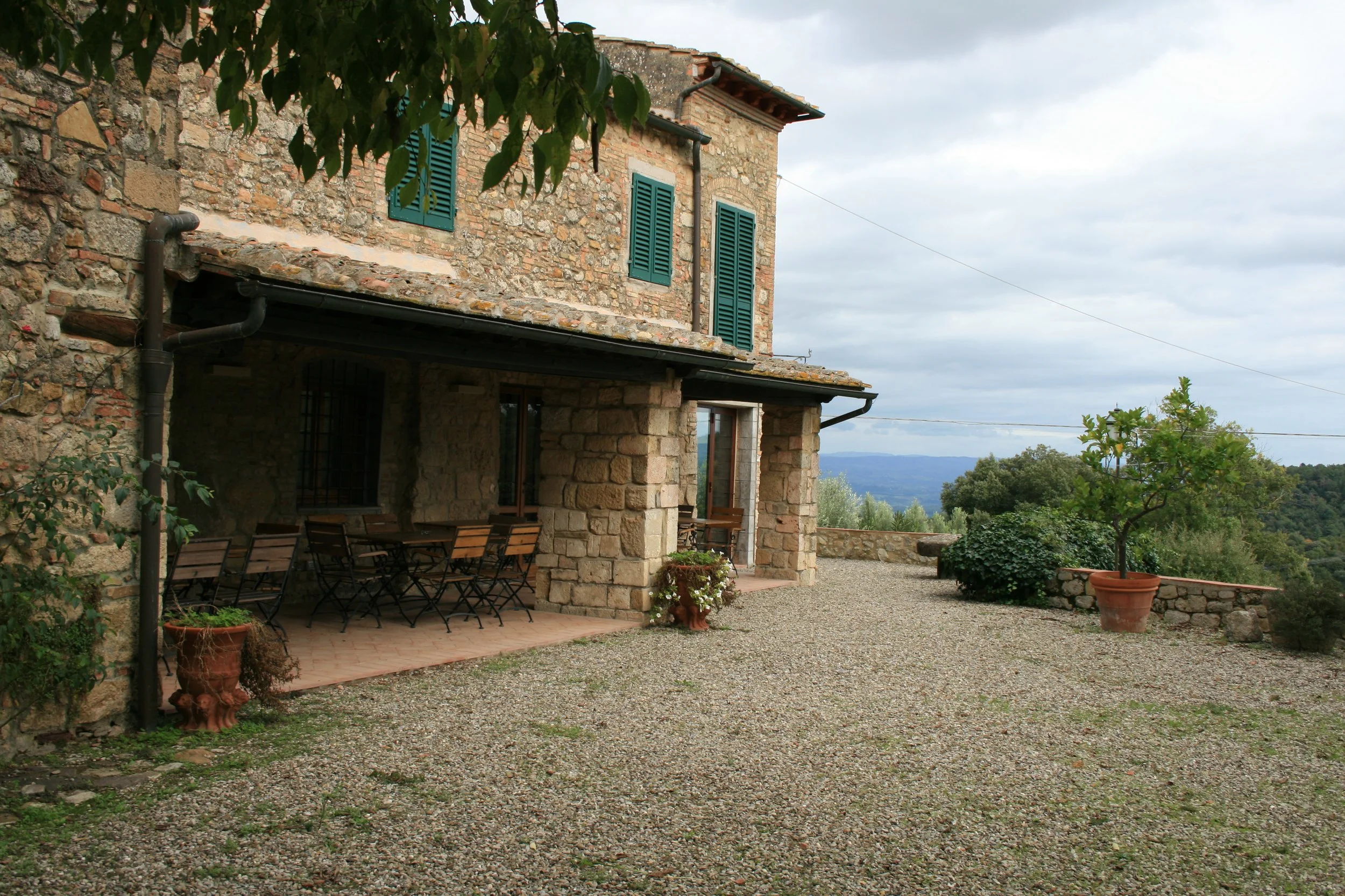 Stone house with green shutters, outdoor patio with wooden table and chairs, potted plants, gravel ground, and scenic countryside view in the background.