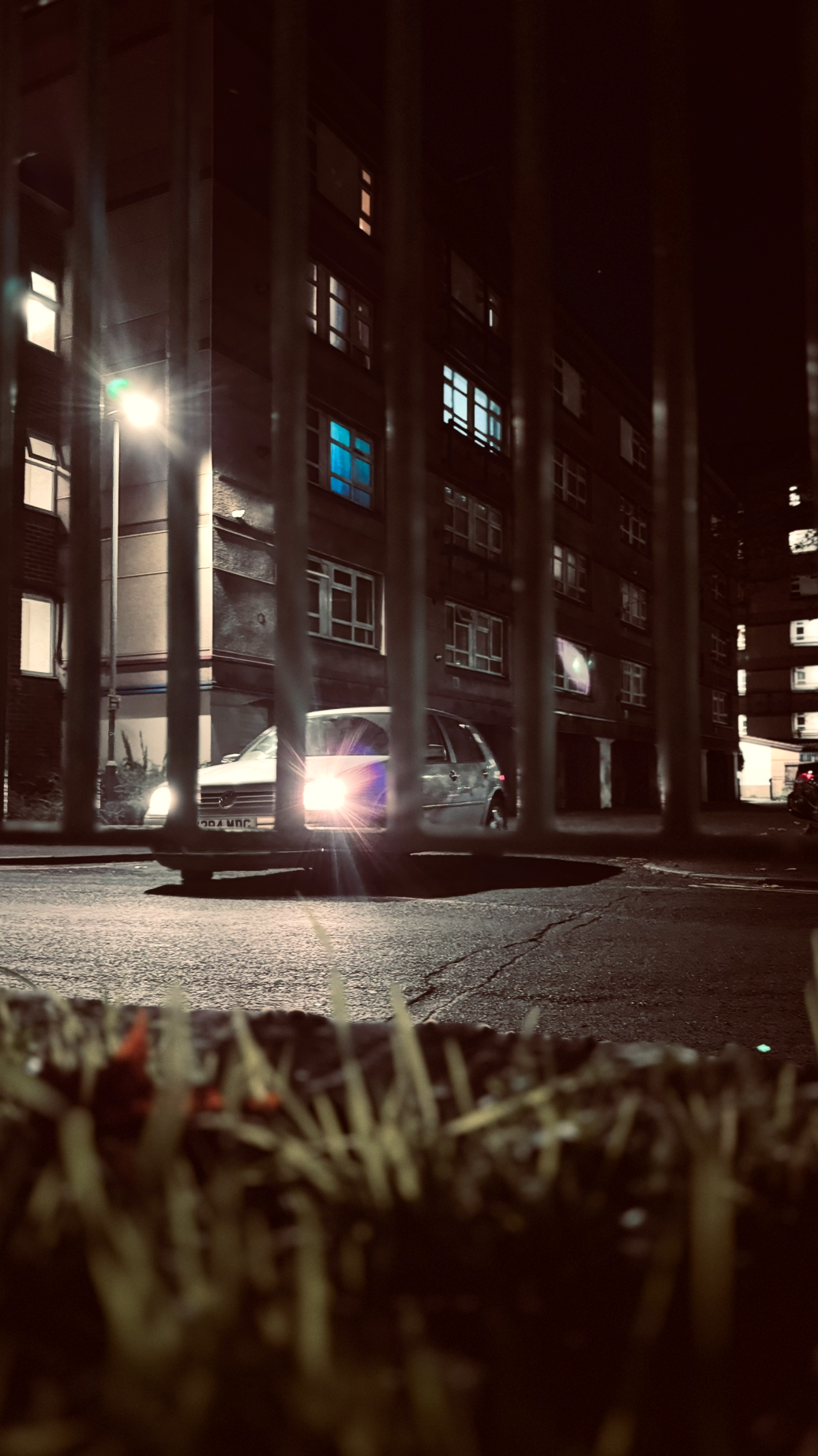 Nighttime view of a residential building parking lot with a car and a parking lot fence in the foreground.