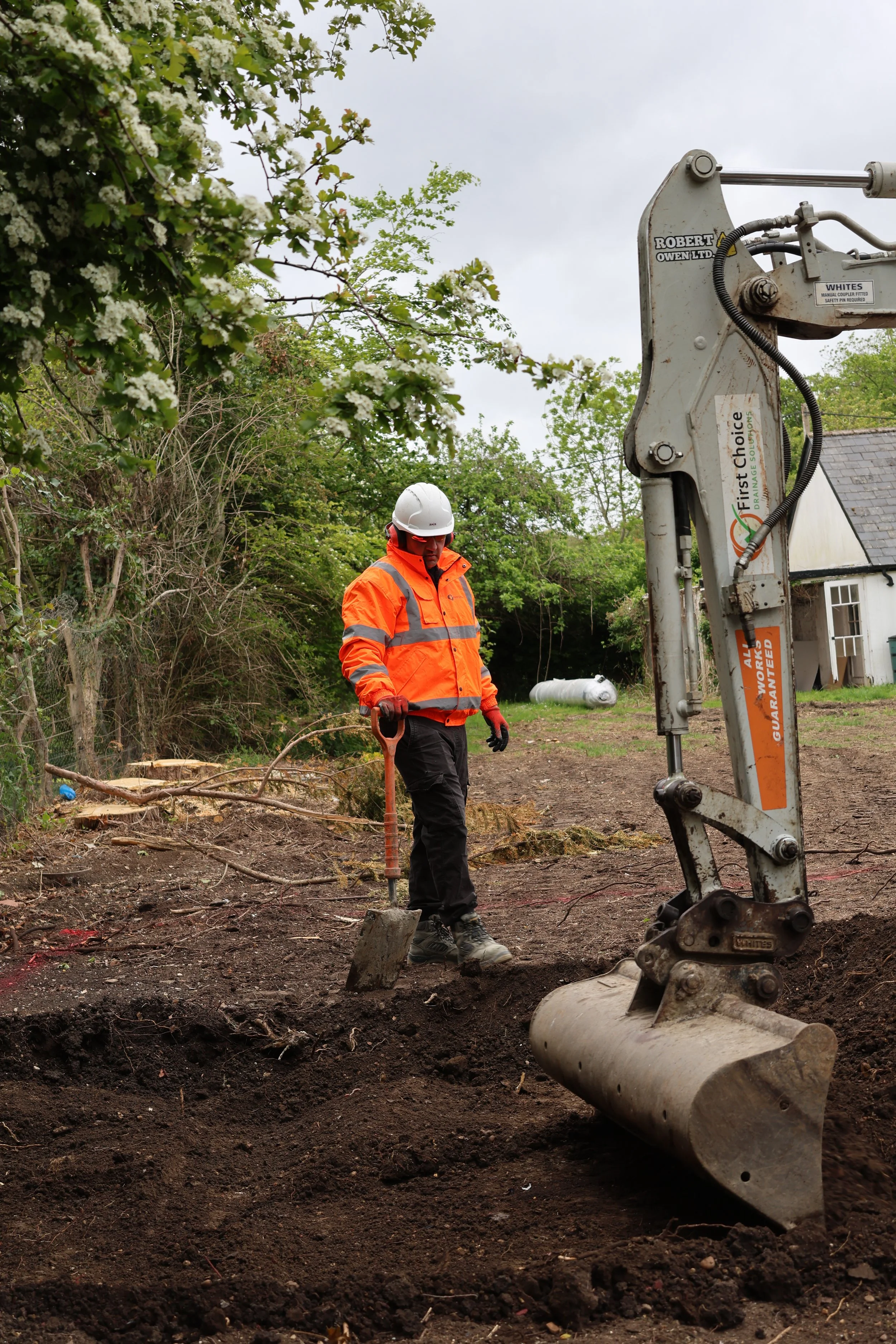 A construction worker in an orange safety jacket and white helmet working with a small excavator on a dirt site. Trees and a house are visible in the background.