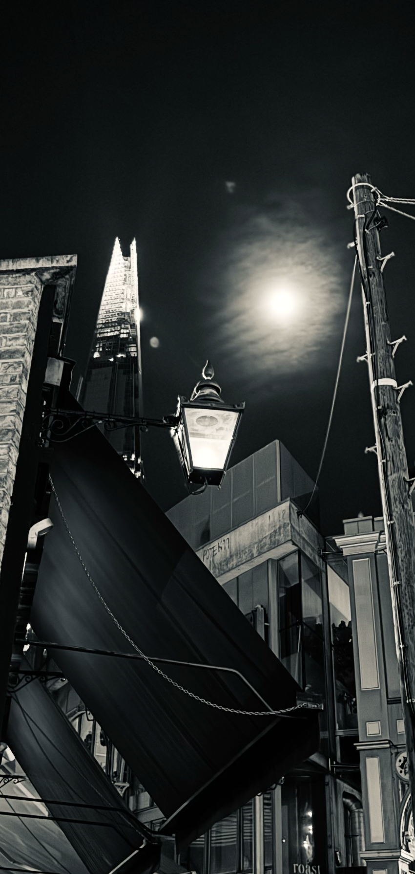 Nighttime city scene with a glowing moon, a modern skyscraper, a streetlamp, a brick building, and a utility pole with wires.
