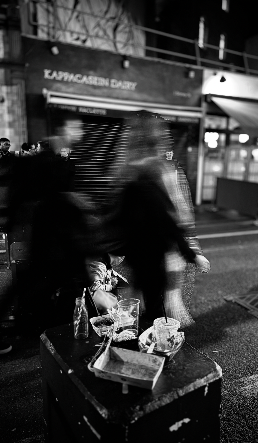 Black and white photo of a city street at night, with blurred pedestrians walking past an outdoor table with used food containers and drinks, and a closed storefront in the background.