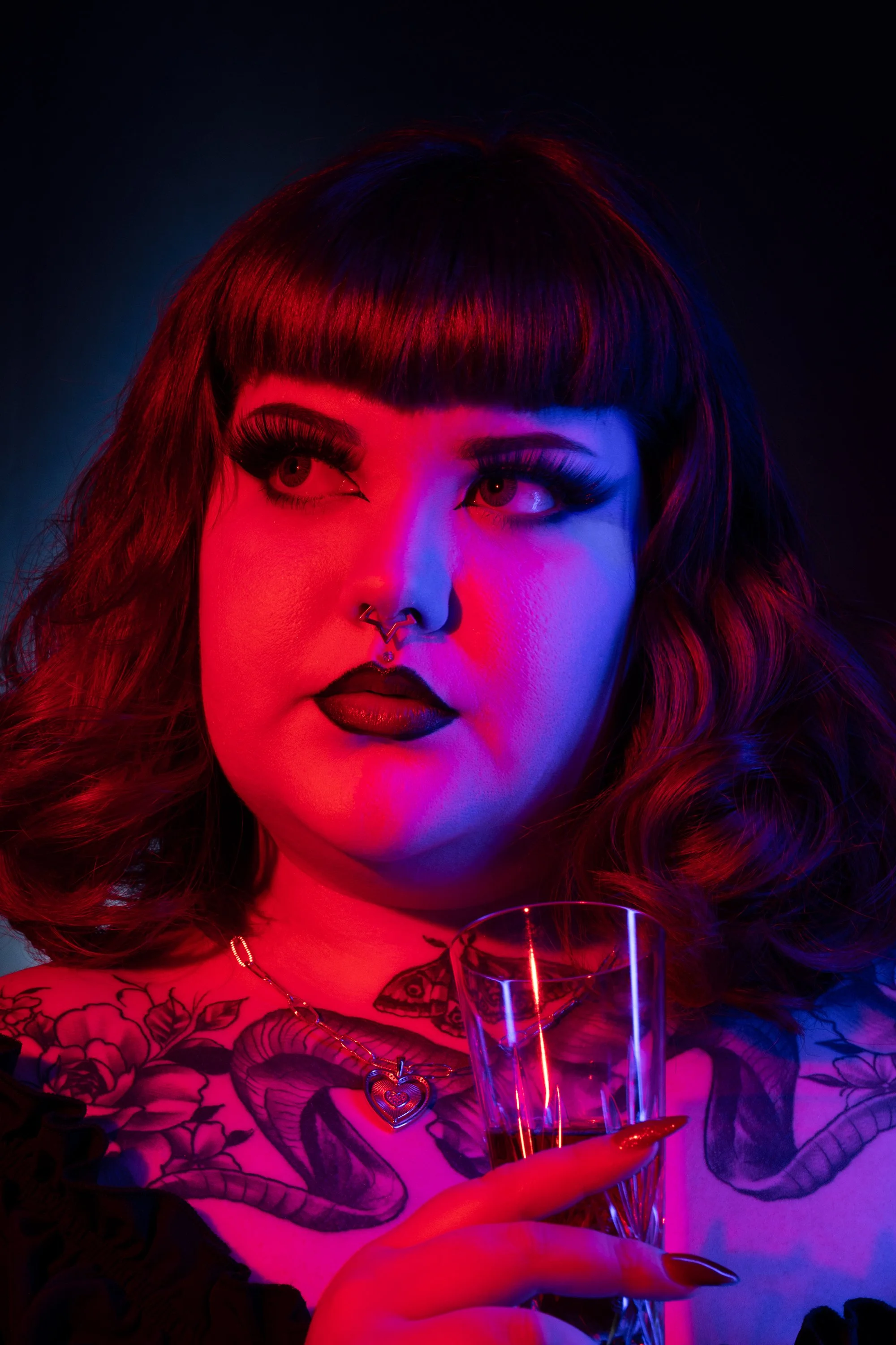A woman with dark lipstick, long lashes, and tattoos on her chest holding a glass of drink, illuminated by colorful red and blue lighting.