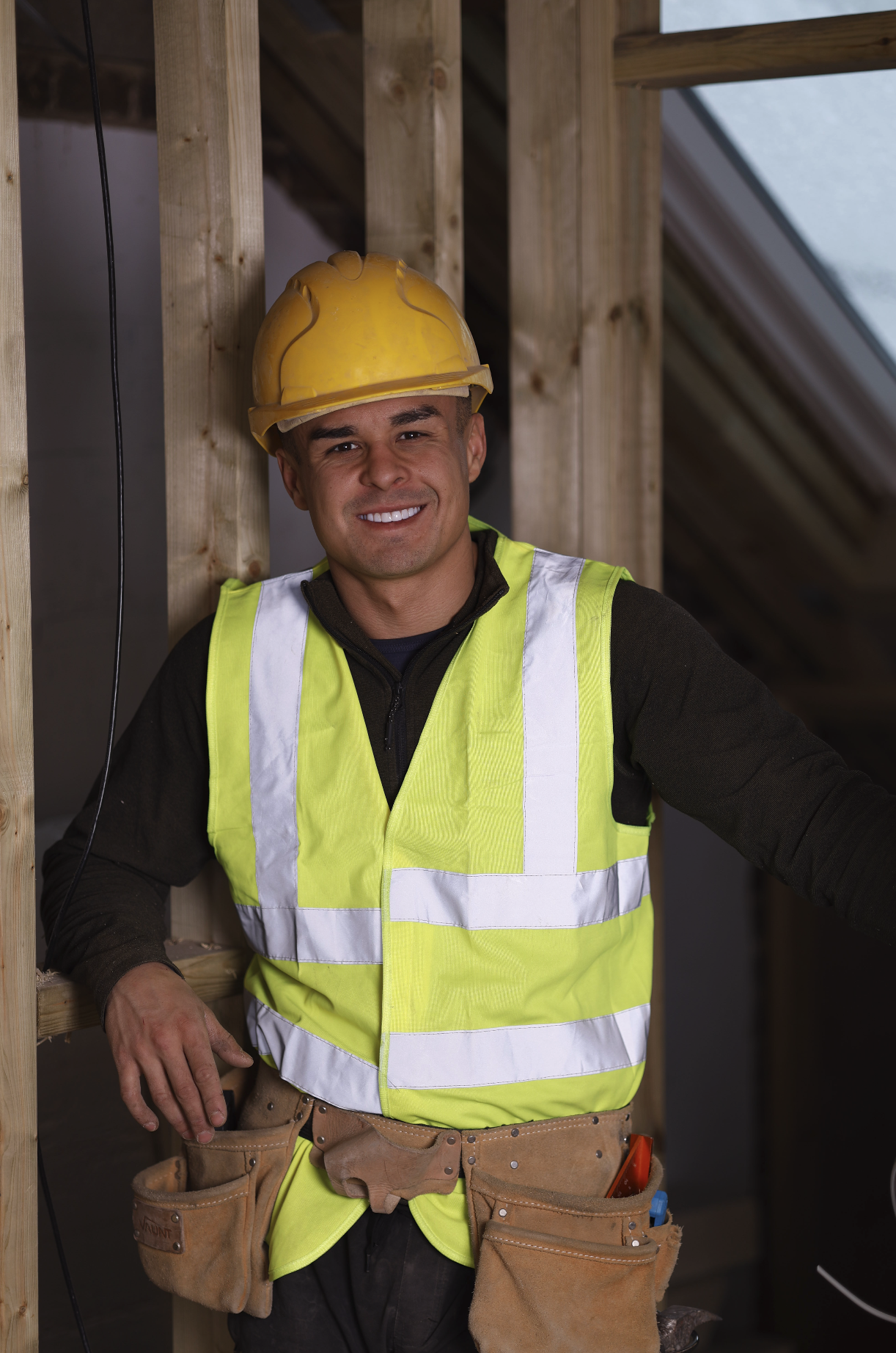 A smiling construction worker wearing a yellow hard hat and high-visibility safety vest, standing inside a building under construction.