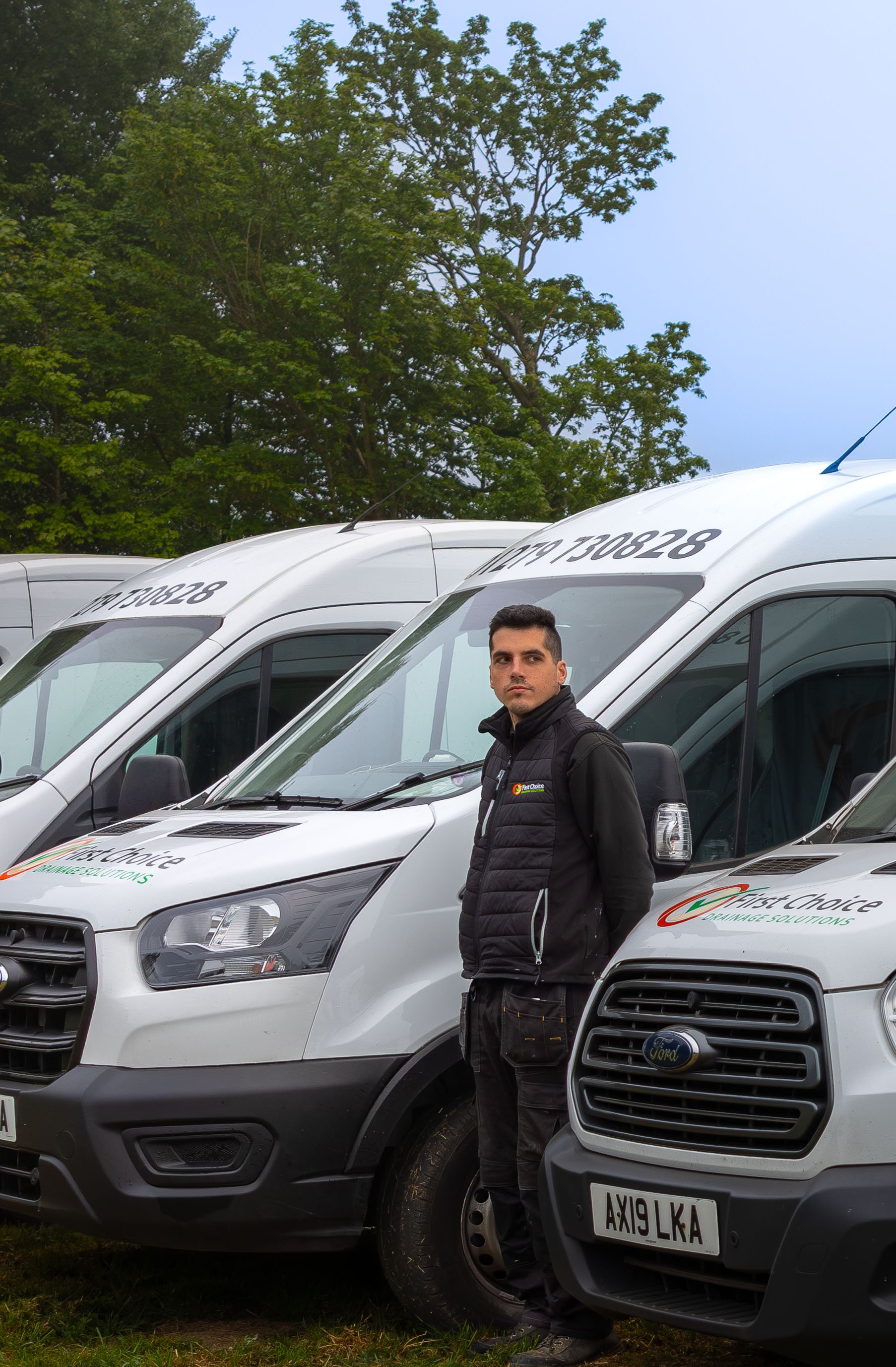 A line of white work vans parked on grass with a man standing beside one of the vans, in front of green trees, wearing a black jacket and pants.